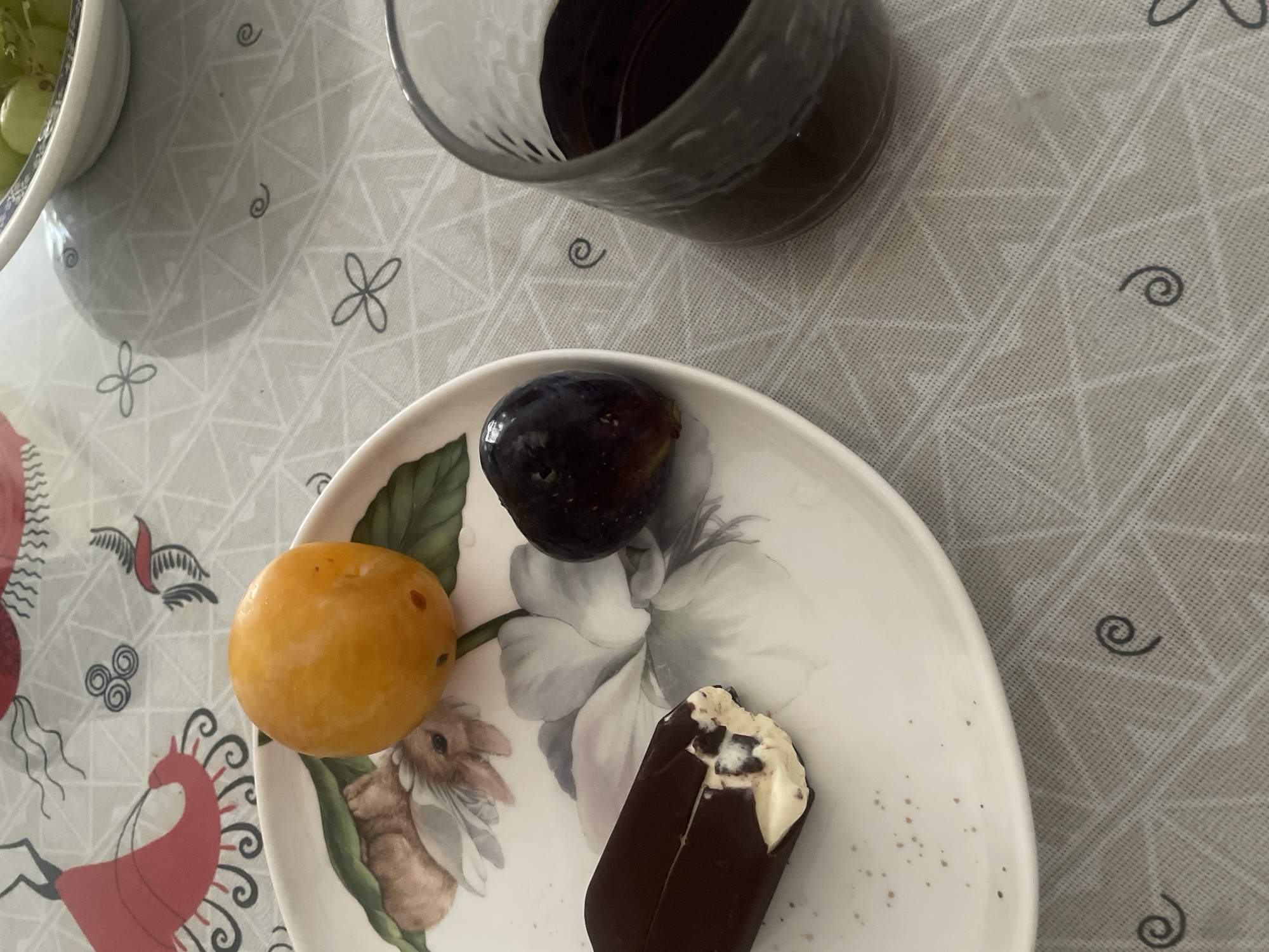 A white plate with a floral design holds a chocolate-covered ice cream bar that has been bitten into, a dark purple fig, and a bright orange plum. To the right of the plate is a dark glass filled with a reddish-brown liquid, likely wine or juice. The setting appears to be an indoor table covered with a light gray tablecloth featuring a subtle geometric pattern and small black swirl designs. The image is taken from a high-angle perspective. The location in Atarfe, Spain, is not discernible from the visual information provided in the image.