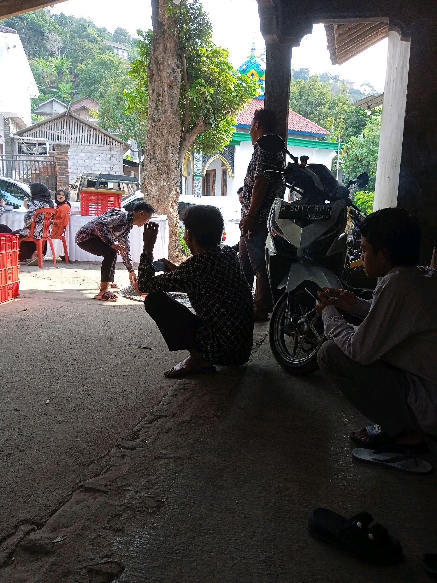 The image depicts an outdoor scene, possibly in a village or rural area, with a mosque visible in the background. Several people are present, engaged in various activities. In the foreground, two men are crouching on the ground. One is wearing a patterned shirt and dark pants, and appears to be looking at something in his hands, possibly a phone. The other man, also in a patterned shirt and dark pants, is looking away from the camera, with his hands near his face.
To the left, a woman in an orange headscarf and patterned top is seated on a red plastic chair. Next to her, another woman in a black headscarf and patterned top is also seated. In the middle ground, a man in a patterned shirt is bending down, reaching towards a mat on the ground. He is wearing sandals.
A white and silver motorcycle is parked to the right of the frame, with a license plate displaying "H 5017 BHG 08-27". Red plastic crates are visible on the left side of the image, near the seated women. The ground appears to be a mixture of concrete and rougher earth. Trees and buildings, including what looks like a mosque with a colorful dome, form the backdrop. The overall lighting suggests it is daytime, possibly late afternoon, with some areas in shadow.