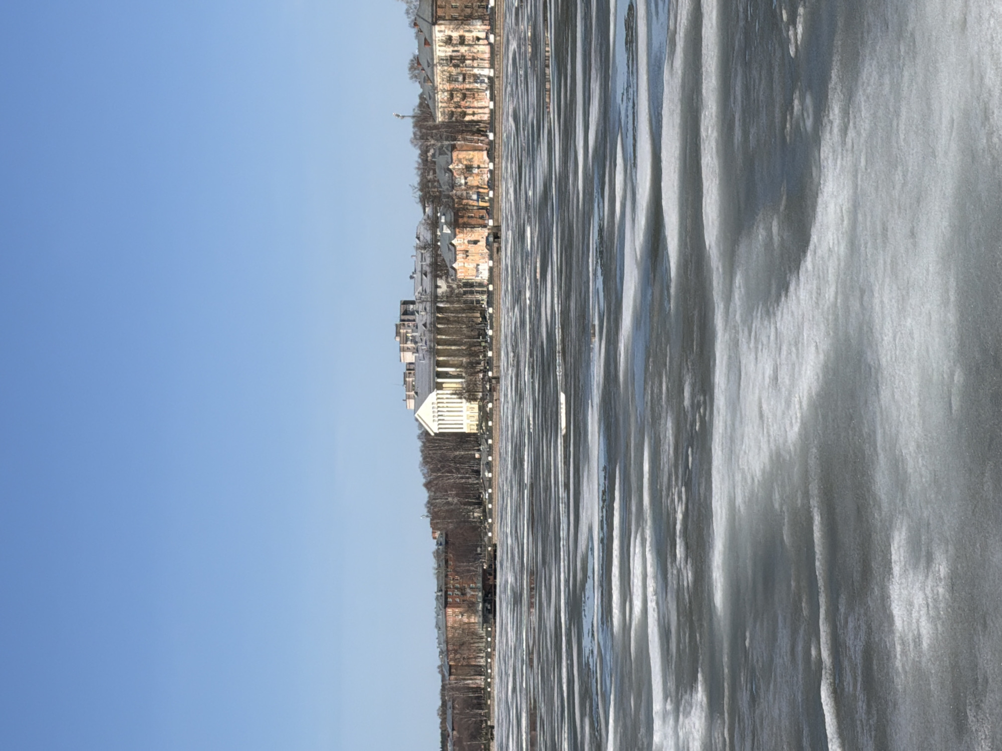An outdoor scene, captured on a sunny day, features a wide body of water with a city skyline on the far bank. The image is oriented to show the horizon vertically, with the sky on the left and the water on the right, as if a horizontal landscape photo was rotated 90 degrees clockwise.
The broad expanse of water in the foreground and midground is covered in a textured layer of ice. This ice displays distinct linear patterns, possibly formed by frozen ripples, broken ice, or refrozen slush, creating alternating bright, reflective strips and darker, more opaque sections. The surface of the ice reflects the clear blue sky and the distant buildings in distorted forms.
On the opposite bank, a cityscape stretches along the horizon. Numerous multi-story buildings are visible, varying in architectural style and color. Some structures have reddish-brown facades, while others appear lighter, including a prominent white building with classical architectural elements. Bare deciduous trees line the water's edge in front of the buildings, their branches devoid of leaves, indicating a winter or early spring setting.
The sky, occupying the left side of the rotated image, is a uniform, clear light blue, suggesting a bright and sunny day, despite the obvious cold weather implied by the ice on the water. No people or specific activities are visible, presenting a serene and still landscape. The scene is located in Votkinsk, Russia. No visible text is present in the image.