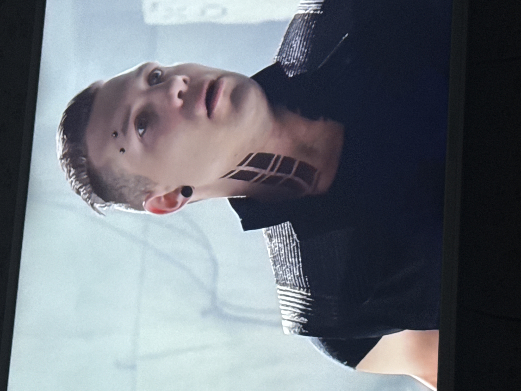 A person with a shaved head and a visible eyebrow piercing is shown from the chest up. They have a grid-like tattoo on their neck and are wearing a dark, button-down shirt. The background appears to be a neutral grey or white color.