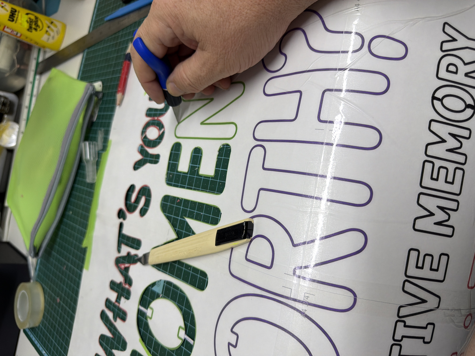The image shows a person using a craft knife to cut out lettering from a large vinyl sheet. The lettering, in various colors, reads "WHAT'S YOUR CREATIVE MEMORY". The person is holding a blue-handled craft knife in their right hand. Another craft knife with a tan handle is nearby on the vinyl sheet. The background includes a green cutting mat, a yellow glue stick, a green pencil case, a metal ruler, and other tools. The location is likely a workshop or crafting area. This image does not provide information on the city of Even Yehuda, Israel.
