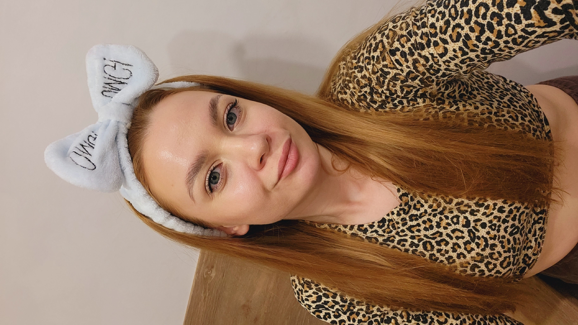 A young woman with long, straight, reddish-blonde hair is positioned in the center of the frame, angled slightly to the left. She is wearing a blue fuzzy headband with bunny ears, inscribed with black lettering. She has fair skin, light eyebrows, and blue eyes. She is wearing a leopard-print long-sleeved shirt that exposes part of her midriff.
The background consists of a plain, light-colored wall on the left and a wooden surface below. The picture appears to be taken indoors, in Chaikovsky, Russia.