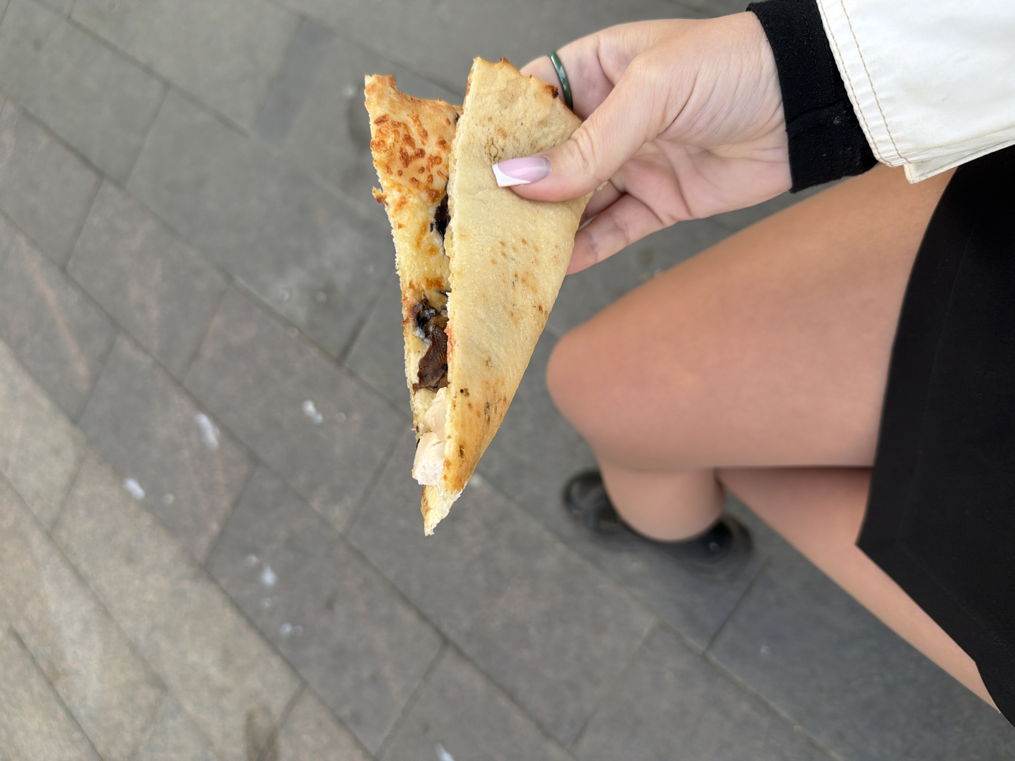 A person stands outdoors on gray brick pavement, holding a folded, triangular crepe or flatbread. The crepe appears to be filled with cheese, mushrooms, and pieces of chicken. The person is wearing a black dress and a light-colored jacket with black trim at the sleeve. The image was taken in a public space in Moscow, Russia.