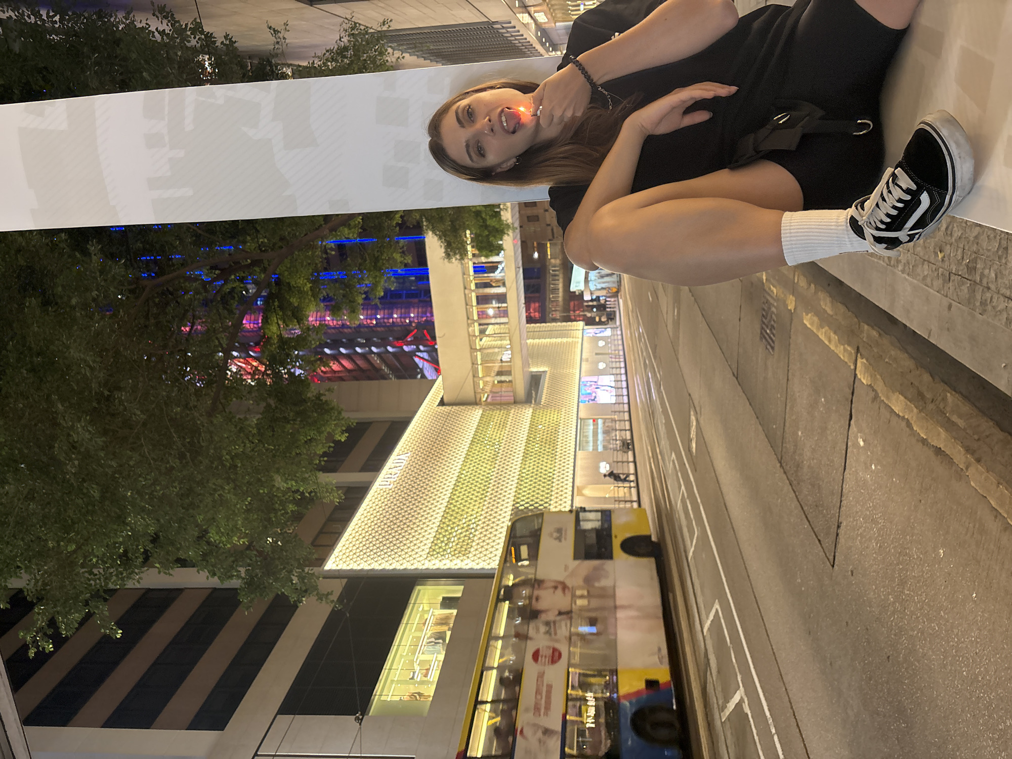 A young woman sits on a ledge in Hong Kong, China. She wears a black outfit with a black belt and black and white sneakers with white socks. She has brown hair and is holding a lighter in her mouth, with the flame showing. Behind her are buildings, including one that is brightly lit and labeled "PRADA," a tree, and a double-decker bus. The street appears to be a paved road.