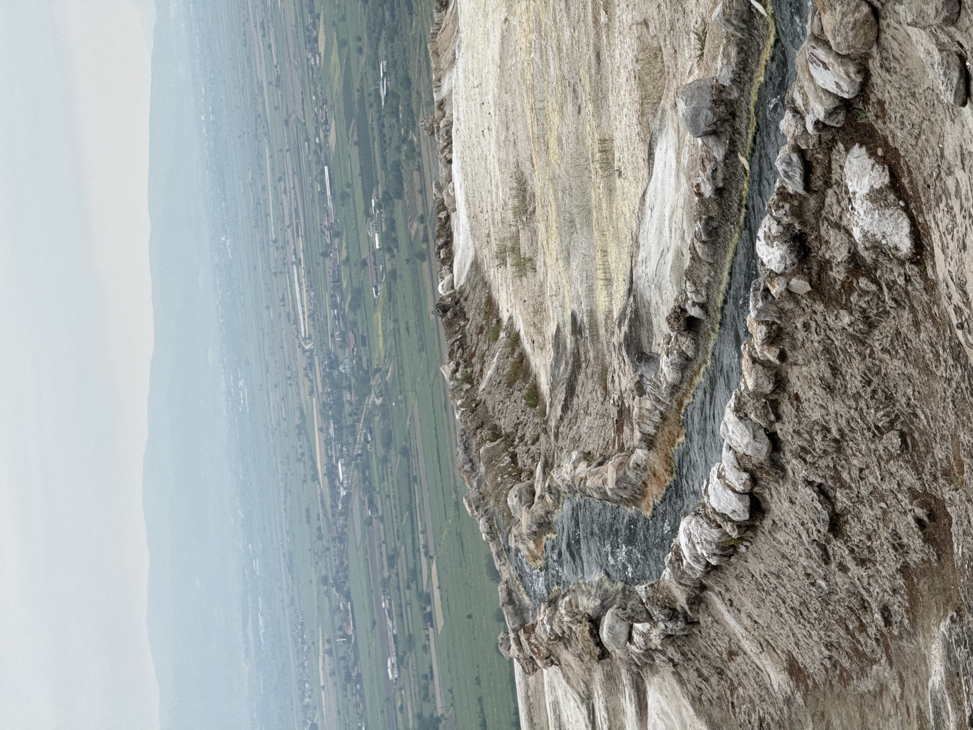 The image presents a view from the travertines of Pamukkale, Turkey. The landscape includes a rocky, cliff-like formation with mineral deposits, likely calcium carbonate, creating a tiered, white appearance. Water flows down the side of the structure, creating a dark streak amidst the pale surface. In the background, a valley is visible, with fields and scattered buildings. The sky is overcast. There are no people in the frame.