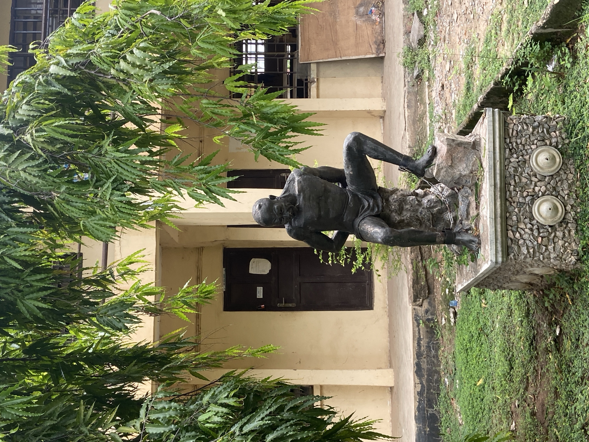 The image shows a bronze statue of a seated man, likely a historical figure. The statue is positioned on a stone base, which has a pebble-dash finish and two circular protrusions, possibly faucets or decorations. The location is an exterior courtyard or garden in Lokoja, Nigeria. A building with a beige facade, dark wooden doors, and windows with metal bars is visible in the background. A large tree with long, narrow leaves partly obscures the view of the building and statue. The ground is covered in grass and some weeds. There's some visible debris near the statue base, which implies the site may not be perfectly maintained. The scene is captured in daylight.