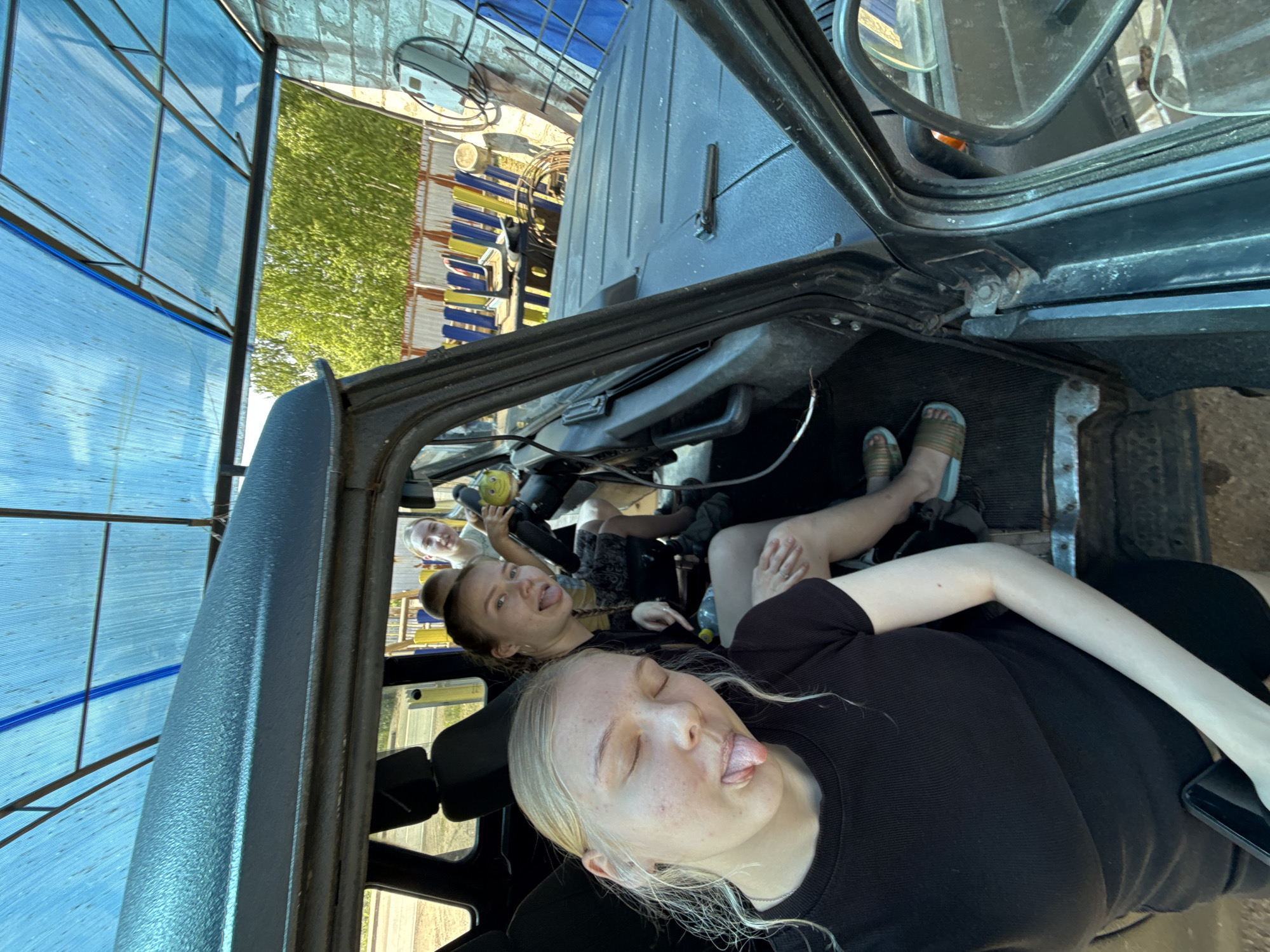 Three young women are inside a vehicle. One woman in the foreground has blonde hair, a black shirt, and her tongue is sticking out. Another woman in the midground also has her tongue sticking out and is holding a yellow object. A third woman is visible in the background. The vehicle has a blue tarp overhead and appears to be parked near a building with yellow and blue objects. The image was taken in Zhigalki, Russia.