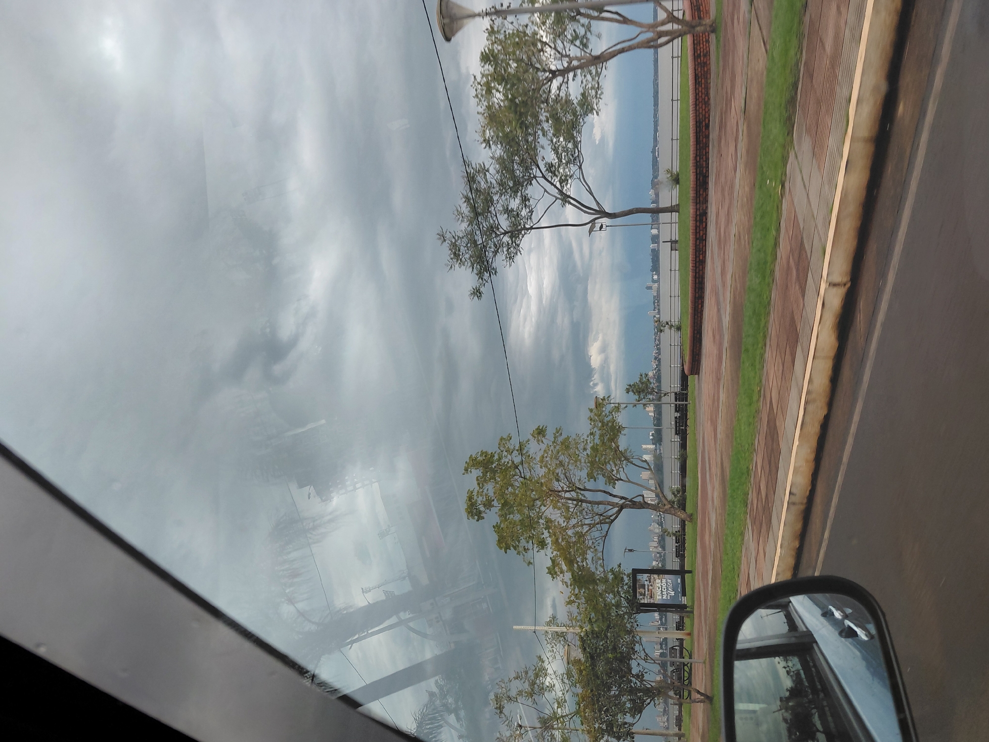 The image captures a cloudy sky over the waterfront in Encarnación, Paraguay. In the foreground, a portion of a car's interior is visible, with the driver's side mirror reflecting the scene. The reflection shows a street, a sidewalk with trees, and a body of water. The main view encompasses a paved walkway bordered by grass and a low brick wall, leading towards a wide river or bay. Across the water, the cityscape of Encarnación is discernible with its buildings and skyline. Several trees are planted along the waterfront promenade, some with sparse foliage. A streetlamp stands near one of the trees. In the distance, a billboard with text is positioned near the water. The overall atmosphere suggests an overcast day with potential for rain.