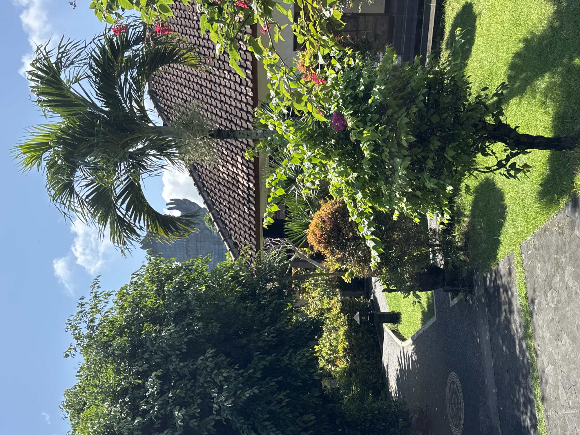 The image captures a sunny outdoor scene in Kuta Selatan, Indonesia, dominated by lush greenery and architectural elements. A palm tree stands prominently against a backdrop of a tiled roof, possibly of a residential building. Other trees and shrubs, some with colorful flowers, fill the foreground and midground. A paved pathway runs along the side, and a glimpse of a distant structure, possibly a building, is visible through the trees under a blue sky with scattered clouds. The scene suggests a well-maintained garden or outdoor space in a tropical setting.