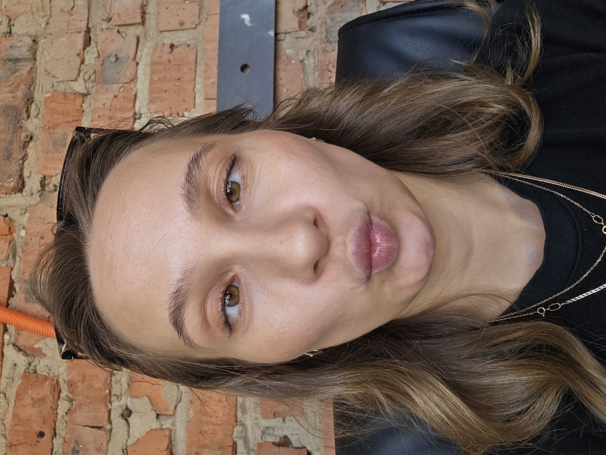 A medium close-up captures a woman with light brown hair and fair skin. Her eyes are hazel, and her lips are slightly puckered. She is wearing a black top and a delicate gold necklace. Black-framed glasses are positioned on the left side of her head.
The background consists of a red brick wall, a dark gray vertical surface, and what appears to be a black leather chair.
The image provides no information to confirm a specific location in Chaikovsky, Russia.