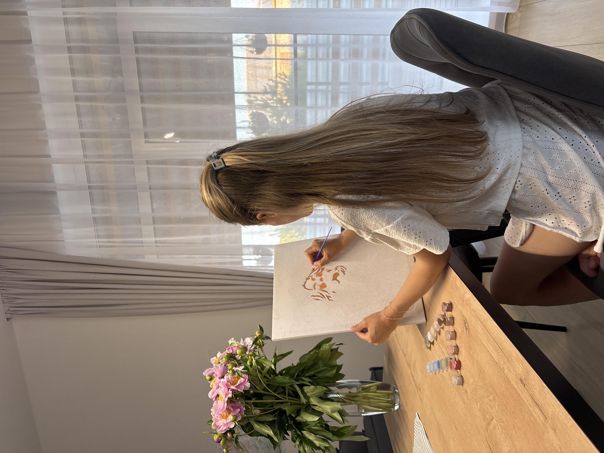 A young woman with long blonde hair, pulled back with a square clip, sits at a wooden table, painting on a canvas. She is wearing a white eyelet top and shorts. Her right hand holds a paintbrush, and she is applying paint to a partially completed image of a face on the canvas. To her left, on the table, are several small containers of paint. In the background, a vase of pink peonies sits to the left of the canvas. The scene appears to be indoors, with a window and sheer curtains behind the woman. The overall environment suggests a home setting. This image was taken in Chaikovsky, Russia.