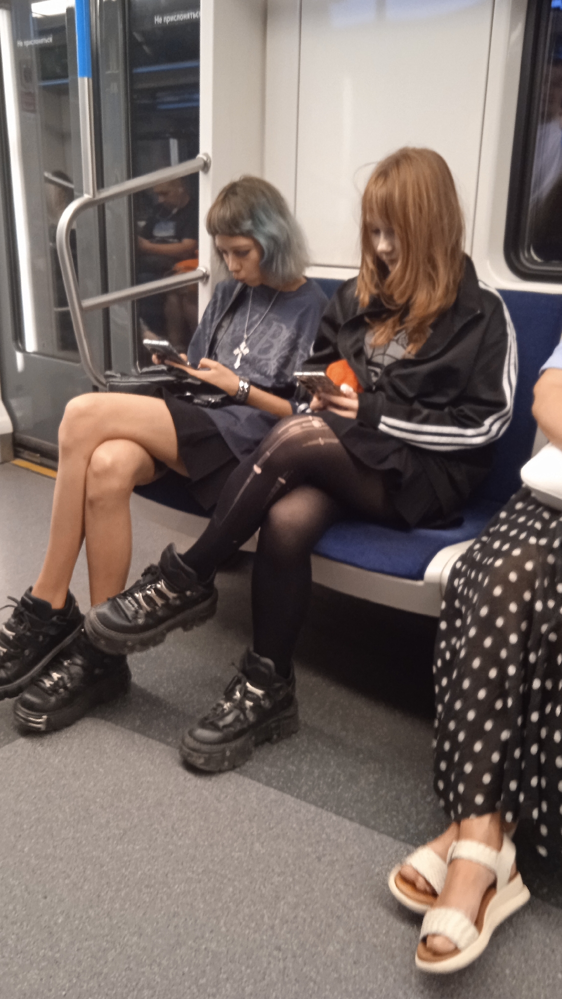 Two young women are seated next to each other on a subway car. The woman on the left has short, blue-tinted hair and is wearing a dark t-shirt with a silver cross pendant, a black skirt, and black platform sneakers. She is looking down at her phone. The woman on the right has long, reddish-brown hair and is wearing a black zip-up jacket with white stripes, a black skirt, ripped black tights, and black platform shoes. She is also looking down at her phone. The subway car has blue seats and a metal handrail. The floor is gray and speckled.