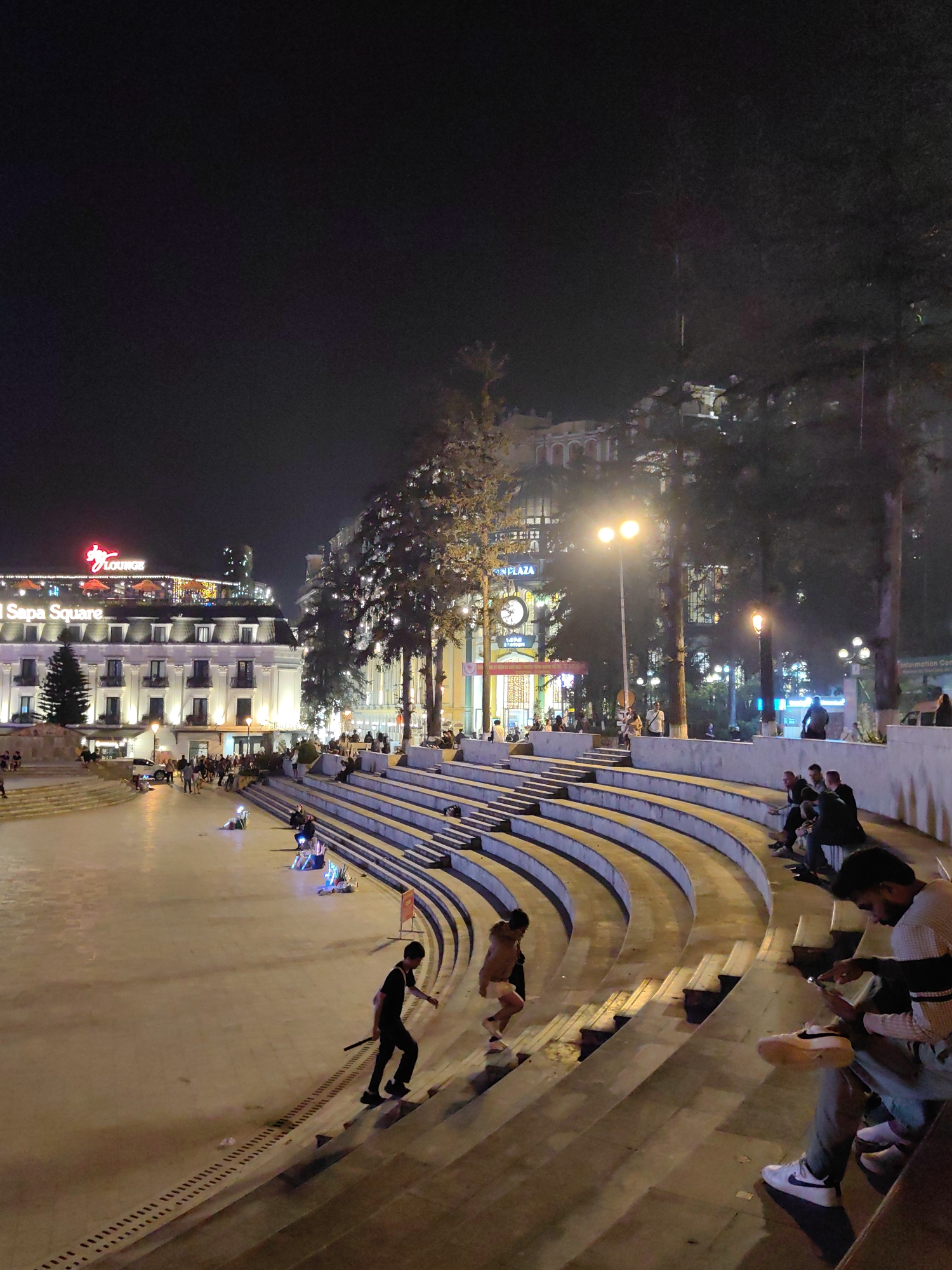 This image depicts a nighttime scene at Sa Pa Square in Sa Pa, Vietnam, viewed from an elevated position looking down towards the main plaza. The square features a prominent amphitheater-style tiered concrete seating area with curved steps, extending from the lower right foreground into the mid-ground.
On the far right in the foreground, a man wearing a striped shirt and light pants is seated on a step, engrossed with a smartphone in his hands. Further left on the steps, another man in a dark t-shirt and dark pants ascends the tiers, holding a dark, stick-like object in his right hand. To his left and slightly higher, a person in light-colored attire walks up the steps. Scattered across the tiered seating, several individuals are visible, some seated and others walking.
The central open plaza floor is illuminated, with a few individuals sitting or lying down, some accompanied by small, glowing objects. In the mid-ground, a crowd of people gathers on the plaza level near a multi-story white building on the left. This building, labeled "Hotel Sa Pa Square" and "Y Lounge" with red illuminated signage, has multiple balconies and lit windows.
To the right of the hotel, behind the tiered seating, is another building with "S Plaza" signage and a large, prominent clock. Trees adorned with string lights are interspersed throughout the area. Various streetlights cast a yellow-orange glow across the square. The background consists of additional illuminated buildings and indistinct foliage, all under a dark night sky.