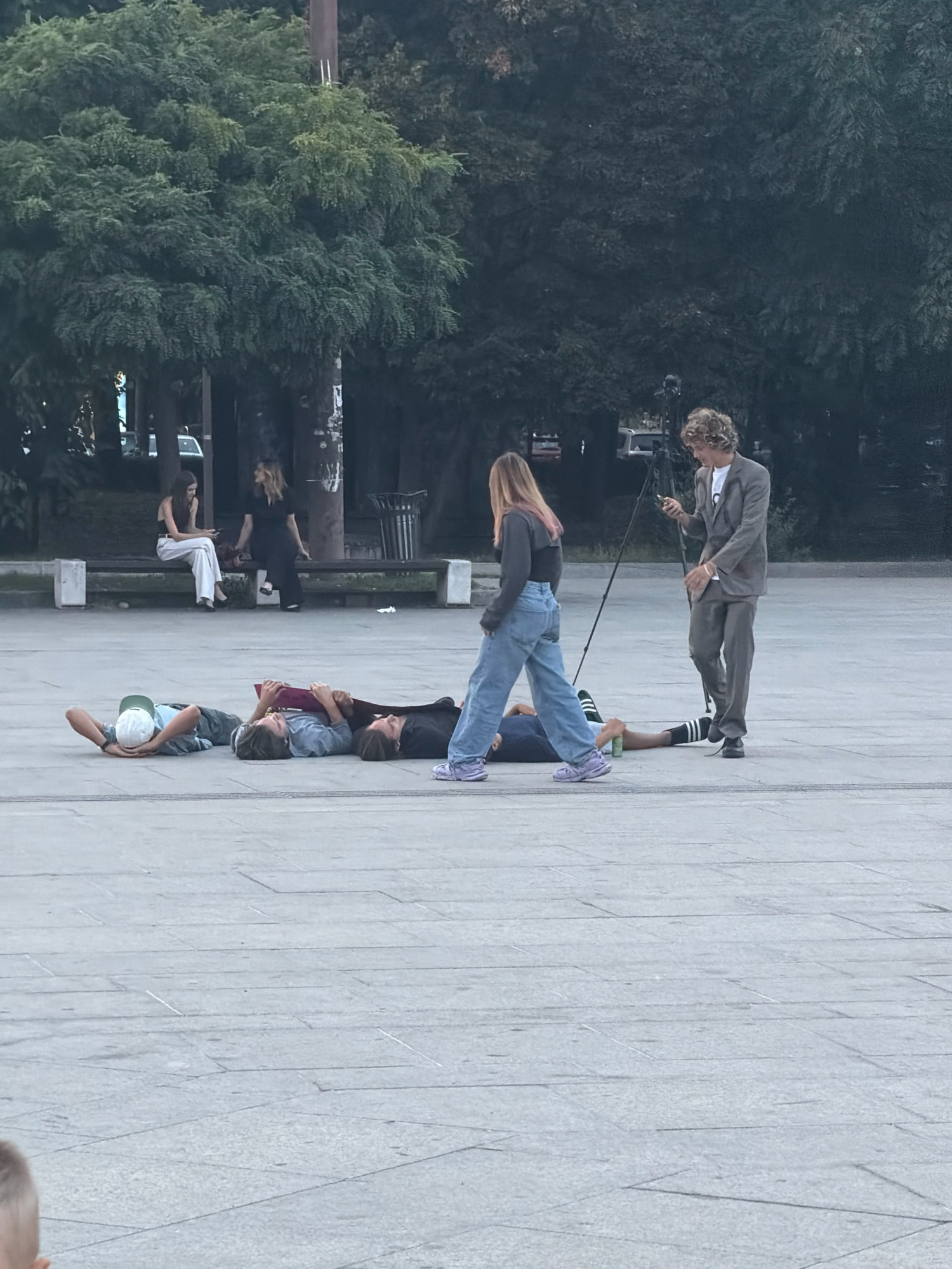 The image depicts a public square in Sofia, Bulgaria, likely near the Ivan Vazov National Theatre, characterized by its paved ground and mature trees in the background. In the foreground, four individuals are lying on the ground, appearing to be resting or posing. Three of them are clustered together, one with a green cap and resting his head on his hands, another with a maroon book, and the third partially obscured. The fourth person is further to the right, with only their legs visible.
In the middle ground, a woman with long blonde hair, wearing a dark top and light blue jeans, walks across the square, her gaze directed downwards. To her right, a man in a light brown suit and white t-shirt stands, looking at his phone and holding a camera tripod. In the background, on a bench, two women are seated; one in white pants and a black top, and the other in a dark dress. Trees and a trash receptacle are visible behind them, contributing to the urban park-like setting.