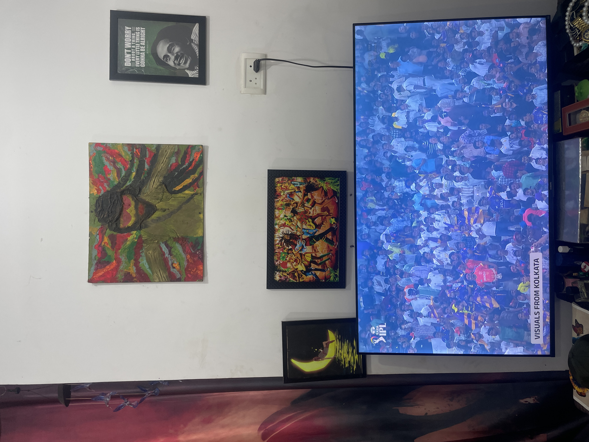 The image shows a wall in an interior setting, with multiple framed pictures and a television set mounted on it. From the text on the TV screen, it is clear the location is Kolkata. The TV is displaying visuals from Kolkata, specifically an event called "TATA IPL". A white wall features a black-framed print with a portrait and the text "DON'T WORRY ABOUT A THING, EVERY LITTLE THING IS GONNA BE ALRIGHT". There is a framed painting with green, red and yellow tones. An electrical outlet and its plug are visible next to the TV. Another framed abstract print is below the outlet, and below it is a print with a figure sitting on the moon. The location is not Morjim, India.
