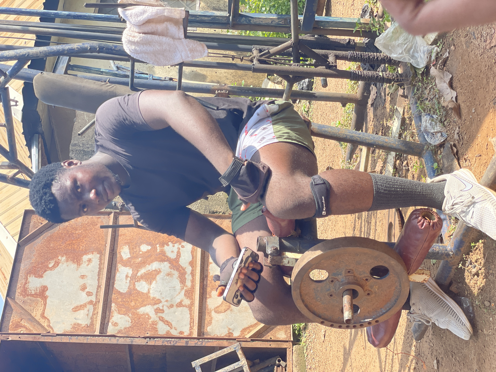 A young Black male wearing a dark t-shirt, olive green shorts over white Nike boxer briefs, grey socks, and grey athletic shoes is seated on a weight machine outdoors. He is wearing black weightlifting gloves and a black knee brace with "Kynclor" written on it. He holds a smartphone in his left hand. The weight machine is made of blue metal and has a large, rusty weight plate on its left side. The background features a rusty metal structure, a towel hanging on a blue metal frame, and some greenery. The ground is reddish-brown dirt. The scene appears to be in a makeshift outdoor gym in Akwanga, Nigeria.