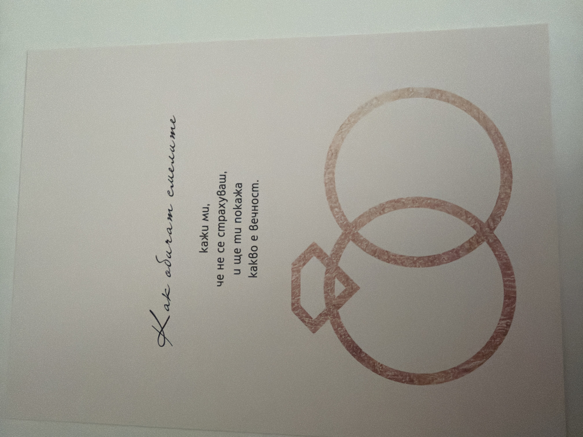 The image is a close-up of a card with text and a drawing of two intertwined rings with a diamond on top. The text is in Bulgarian and appears to be a poem or a romantic message. The drawing is in a light pink, rose gold color. The background is a pale, textured surface. There is no indication of any specific location in Pazardzhik, Bulgaria, as the image is a close-up of a static object.