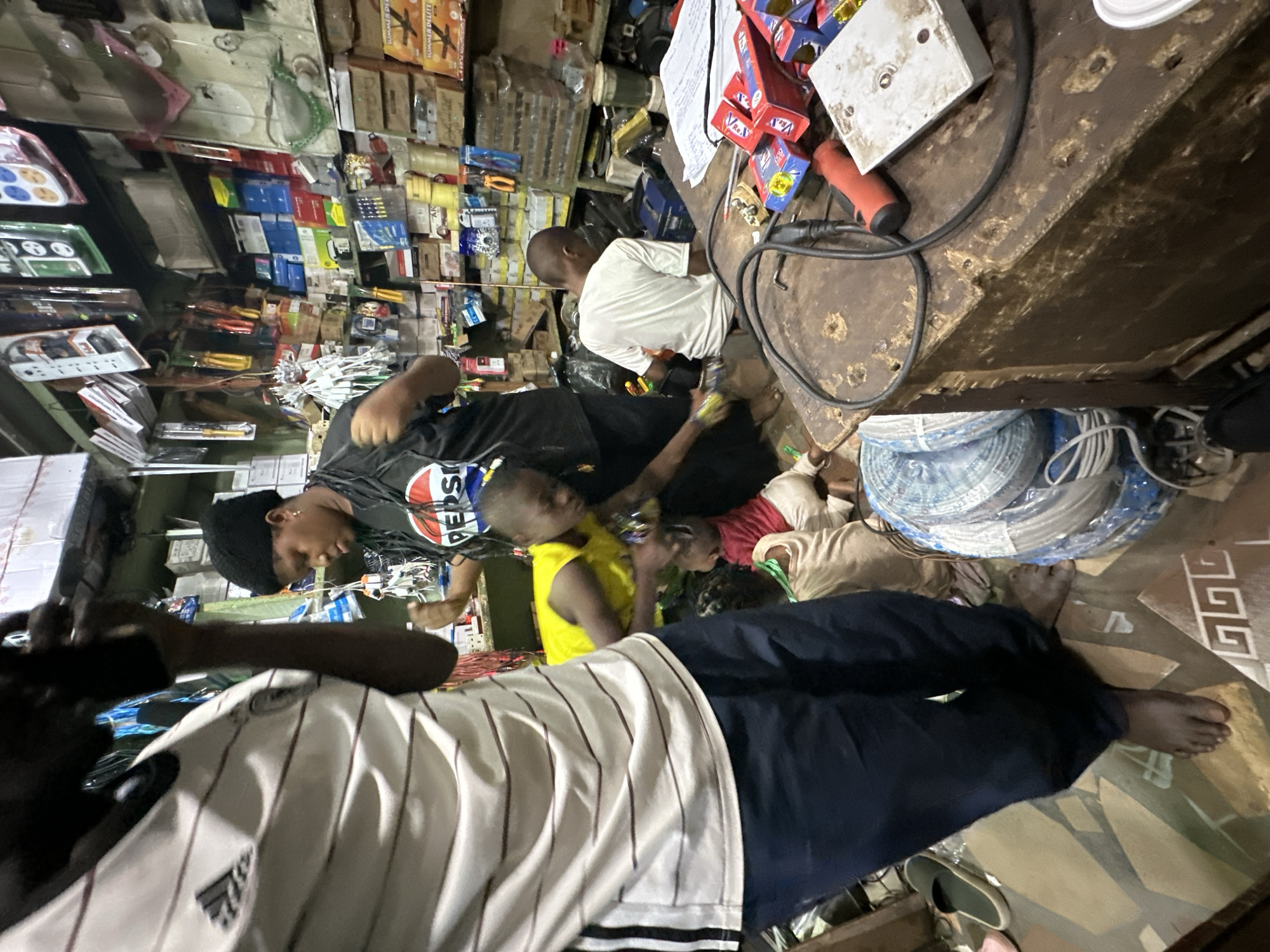 In Barden Yana, Nigeria, a group of individuals are gathered inside a cluttered shop filled with various goods. The shop appears to be a hardware or electronics store, with shelves packed with boxes, tools, and electrical components. In the foreground, a person wearing a white and black striped shirt and dark pants is partially visible, facing away from the camera and holding a phone to their ear. To the right, a woman in a black t-shirt with a Pepsi logo is tending to several younger individuals. One child, wearing a yellow vest, is actively engaged with something on a table, while other children are positioned around them. Another person, wearing a white t-shirt, is seated at the table, looking towards the activity. The shop counter is laden with items, including coiled wires, electrical boxes, and tools. The overall impression is one of a busy, everyday scene within a local Nigerian business.