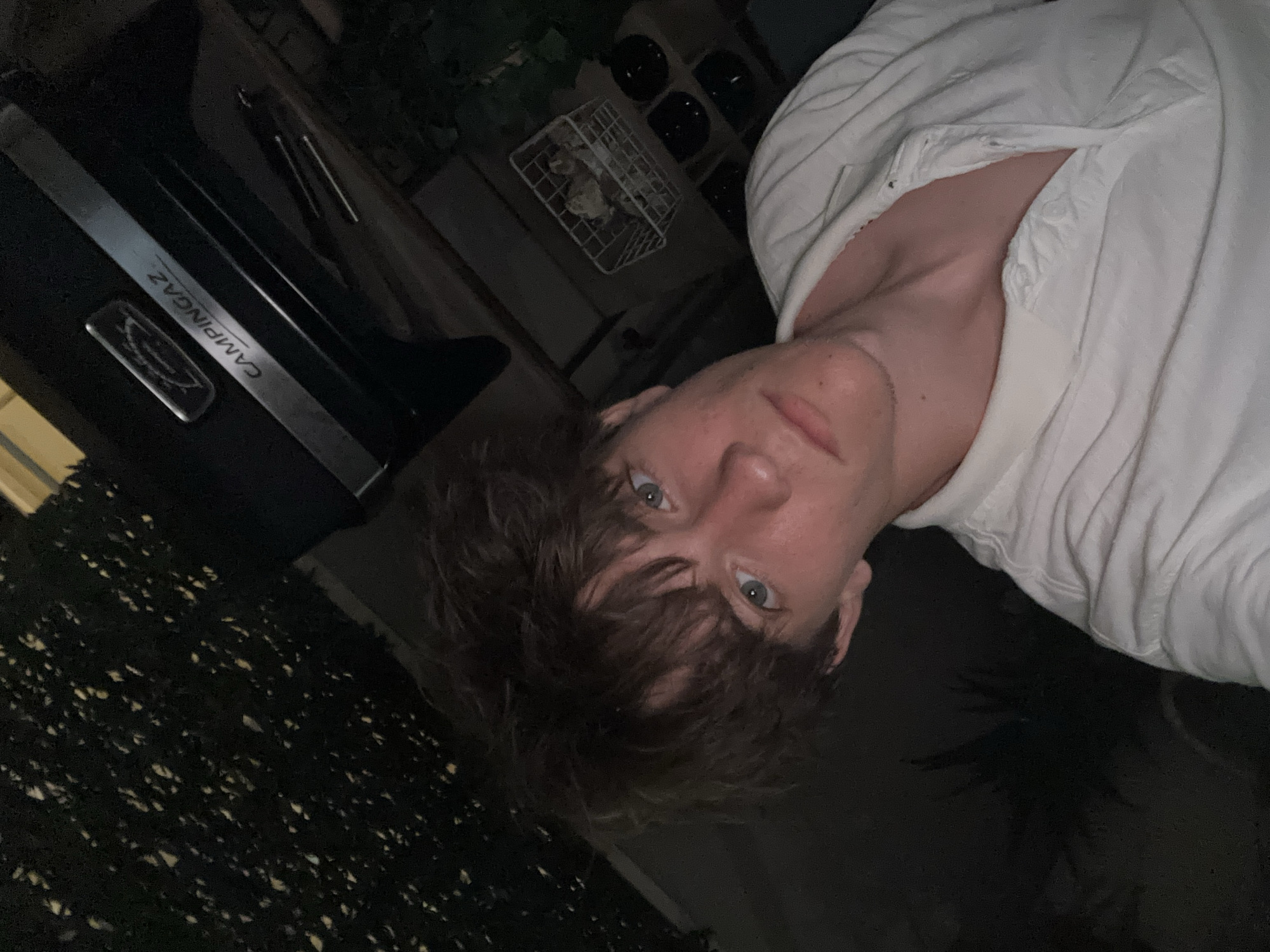 The image shows a young person with light brown hair and blue eyes, wearing a white polo shirt. They are looking directly at the camera with a neutral expression. The background is dark, with some foliage visible on the left and a dark grill with the brand "CAMPINGAZ" on it in the upper right quadrant. There is also a white basket containing items and some black bowls in the background. The scene appears to be outdoors, possibly a backyard or patio area, at night or in low light conditions. There are no discernible landmarks or features that specifically place the image in Cupra Marittima, Italy.
