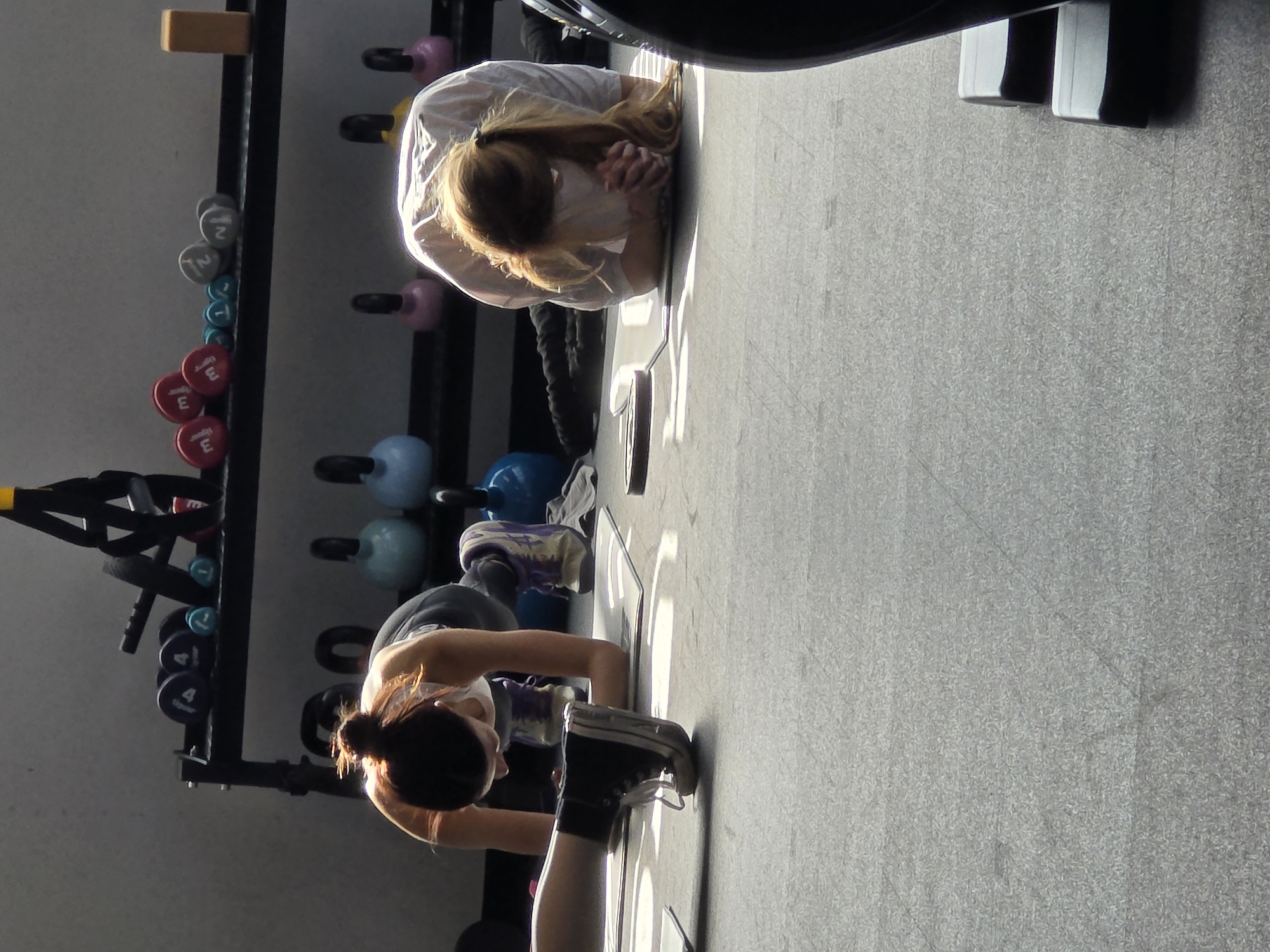 The image, originally rotated 90 degrees clockwise, depicts two women exercising in a gym in Plovdiv, Bulgaria, bathed in strong natural daylight. The floor is covered with a textured grey gym matting, marked with some white rectangular lines.
In the foreground, a woman with dark hair tied in a bun is in a plank or push-up position, facing away from the camera. She is wearing a light-colored top, dark athletic pants, and black sneakers with white laces, actively engaged in her workout.
Further back, another woman with long, lighter-colored hair is lying prone on the floor in a resting or stretching pose, similar to a child's pose. Her head is down, and her arms are clasped in front of her. She wears a white long-sleeved shirt, with the sunlight creating a bright halo around her hair.
To the left of the women, a black metal shelving unit holds various gym equipment. Visible are colorful dumbbells, organized by weight: red ones marked "3" and "1.5" (likely kilograms), grey ones with "2" and "1.25", cyan ones with "1", and blue ones with "4". Below these, several kettlebells in pink, light blue, and yellow are stored. A TRX suspension trainer hangs from the top of the rack, and a small wooden block rests on its uppermost shelf.
The bright, directional sunlight streaming into the gym creates distinct highlights on the women and equipment, casting sharp shadows on the floor. The overall atmosphere suggests a focused and calm fitness environment. No specific emotions are discernible from their obscured faces, but their postures indicate engagement in a workout or recovery session.