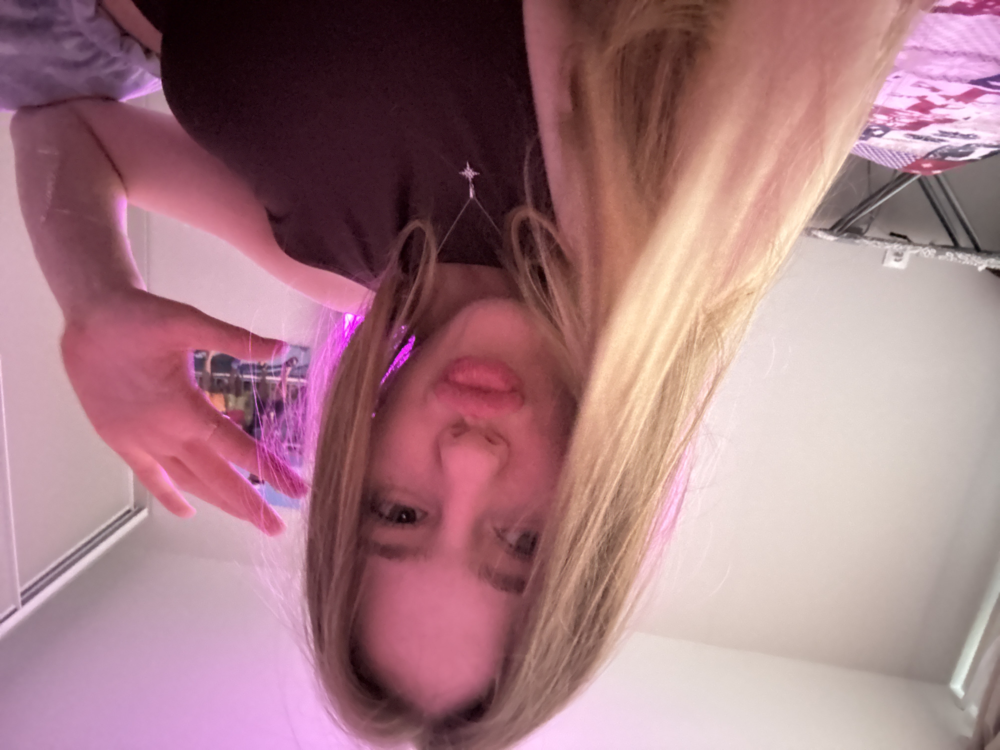 A young person with long, blonde hair is captured in a worm's-eye view, looking directly down at the camera. The subject's face is framed by their hair, with their lips slightly pursed and eyes gazing forward. They are wearing a dark top, from which a delicate silver necklace with a star-shaped pendant hangs. One hand is visible on the left side of the frame, reaching towards the camera or slightly upwards, illuminated by a strong pink/purple light that casts a vibrant hue on the subject's hair and portions of the scene.
The background is an indoor setting, likely a room, characterized by light-colored walls or ceiling. In the upper right corner, a glimpse of patterned fabric, possibly a blanket or duvet cover with red and white designs, is visible, suggesting a bed or living area. Below this, a metallic structure, possibly part of a bed frame or shelf, can be seen. The artificial pink lighting dominates the left side of the image, creating a distinct atmosphere. There is no visible text in the image. The scene appears to be set in Novoe Devyatkino, Russia.