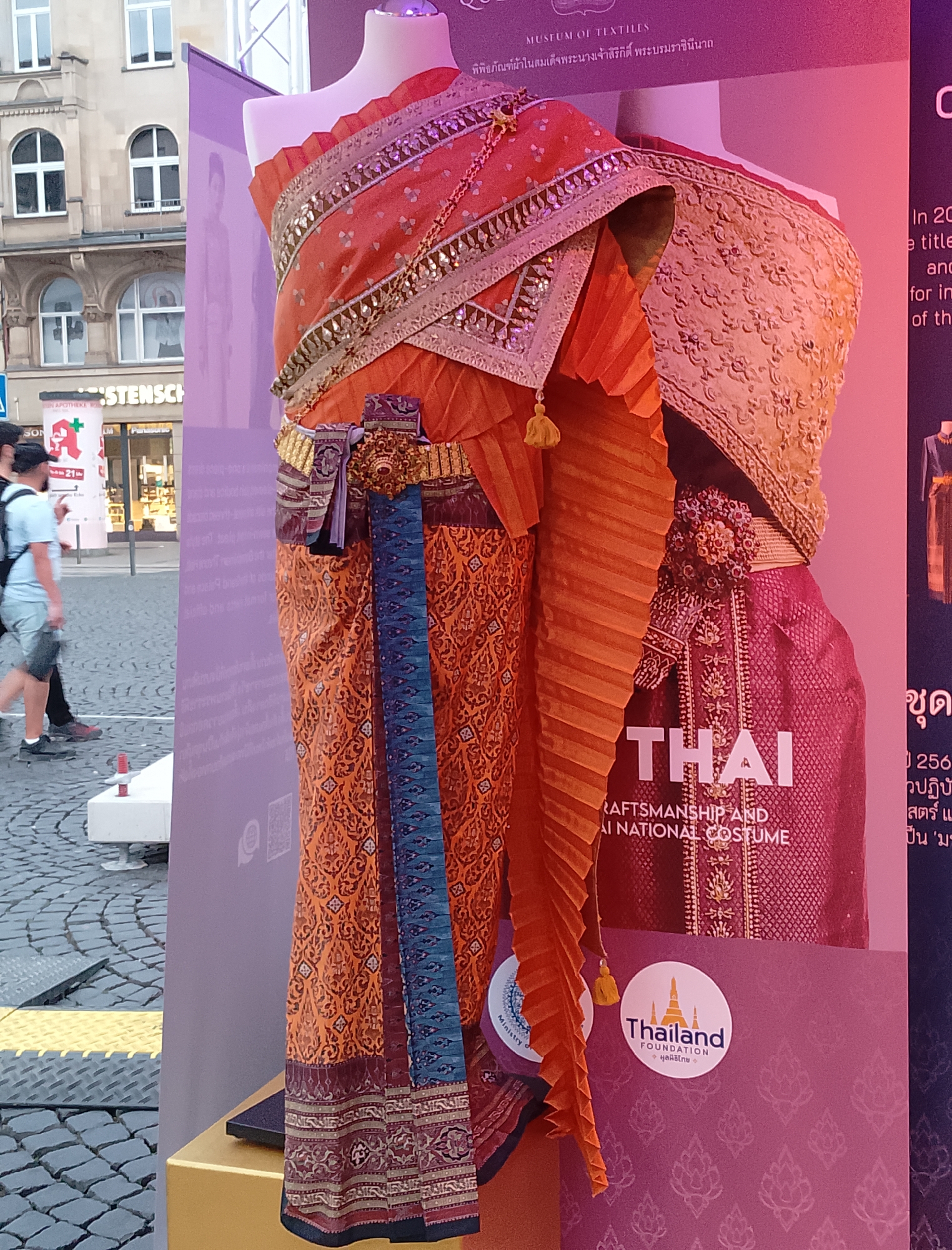 The image shows a display of traditional Thai costumes, likely at an outdoor event in a European city. Two mannequins are dressed in elaborately decorated orange and red silk garments, adorned with intricate gold embroidery, beaded trim, and a decorative belt. The costumes feature pleated fabric panels and flowing skirts. Behind the mannequins, a large purple banner displays text in English and what appears to be Thai script. The English text includes "MUSEUM OF TEXTILES," "THAI CRAFTSMANSHIP AND THAI NATIONAL COSTUME," and a logo for "Thailand FOUNDATION."
To the left of the display, a section of an urban street is visible. Cobblestone pavement is present, along with yellow and black hazard markings. A building with arched windows and traditional architecture stands in the background, suggesting the location is likely in a European city. Two individuals, a man and a woman, are walking on the street, with the man wearing a light blue t-shirt and shorts, and the woman in dark clothing. The overall scene suggests a cultural exhibition or promotional event taking place in a public urban space.