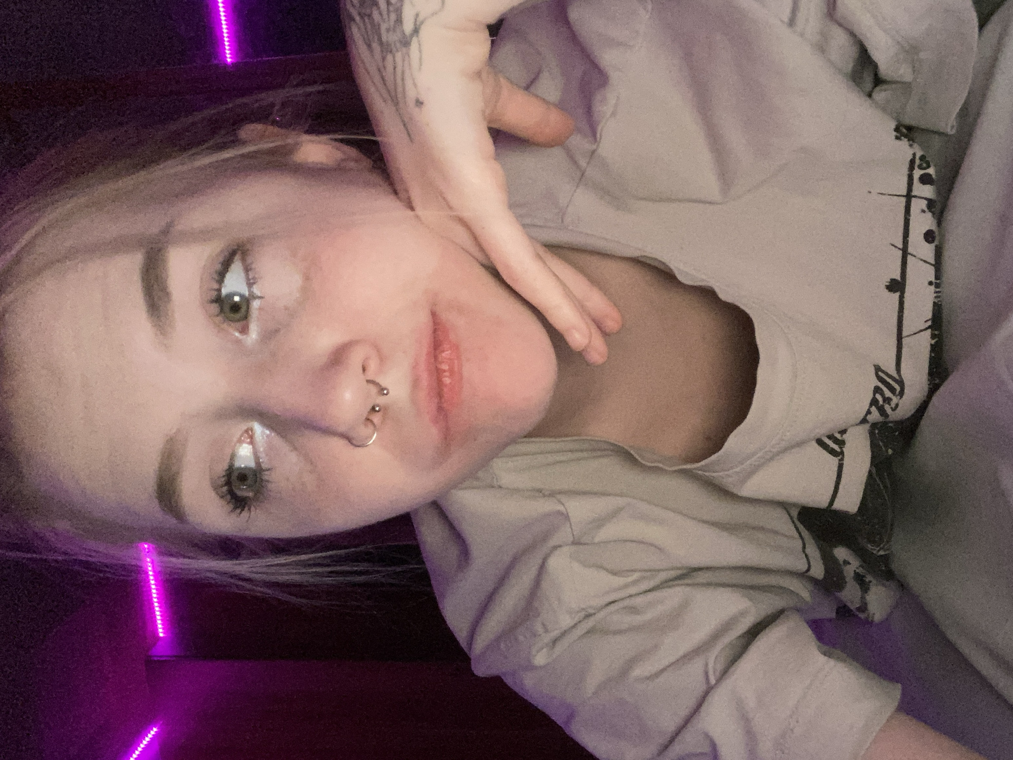 The scene depicts a close-up, medium-shot portrait of an individual, likely female, situated indoors in the city of Izhevsk, Russia. The subject has light-colored hair, green eyes, and a fair complexion. Visible facial features include a septum piercing with two small spheres and a single nostril hoop piercing on the left side of the nose. White eyeliner or shadow is applied to the lower waterline of both eyes. The subject's left hand rests with fingers against the jawline and chin. A faint, dark tattoo is visible on the dorsal side of the subject's left hand, near the wrist. The individual wears a light-colored, possibly beige or light grey, short-sleeved t-shirt with a dark graphic design partially visible across the chest. The background is dimly lit, with a horizontal strip of bright pink LED lights mounted along a dark wall in the upper left quadrant of the frame, casting a magenta hue on the environment and portions of the subject. The subject's gaze is directed upwards and to the right of the frame, suggesting an averted or contemplative expression.