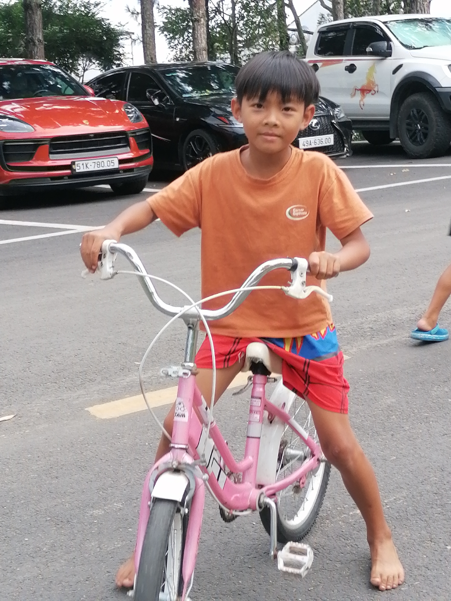 In Da Lat, Vietnam, a young boy with dark hair stands in front of a pink bicycle. He is wearing an orange t-shirt and red shorts with a blue and yellow pattern. The boy is holding the handlebars of the bicycle, which has white grips and a white brake lever. The bicycle has a white fender and a pink frame. In the background, there are several vehicles, including a red Porsche SUV, a black sedan, and a white pickup truck. The ground is asphalt, and there are yellow lines painted on it, indicating a road or parking area. Trees are visible in the background. The overall scene appears to be an outdoor setting, possibly a parking lot or a street in Da Lat.