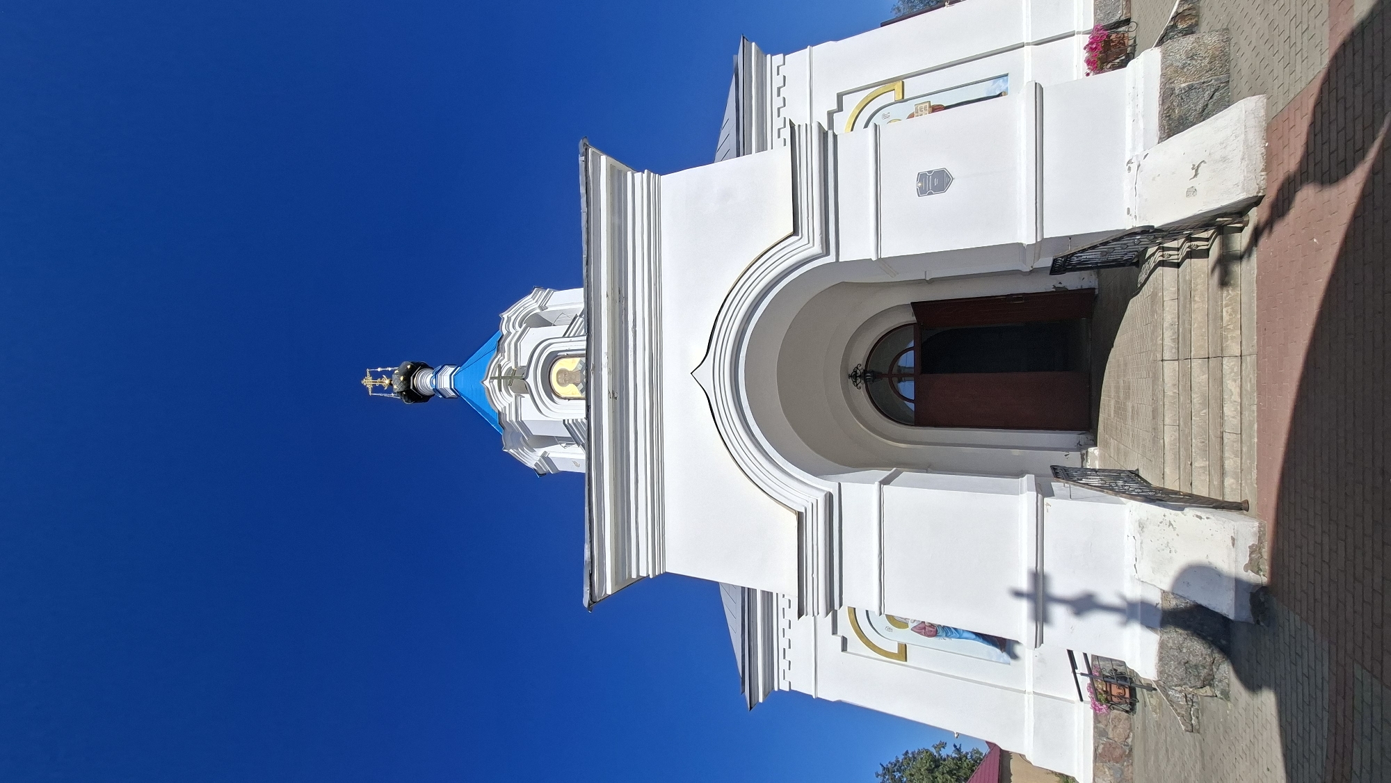 An eye-level, wide-angle, full shot captures the exterior of a white Orthodox church under a clear, bright blue sky. The church features a prominent, arched entrance with dark brown double doors, partially open. Stairs lead up to the entrance, flanked by black metal railings. The building is adorned with architectural details like cornices and decorative elements, and on its facade, there are religious icons. A blue-domed cupola with a golden cross crowns the structure. To the left of the entrance, a shadow of a cross is cast on the white wall, indicating the time of day. The foreground consists of a paved walkway with red brick patterns. In the background, the edge of a red-roofed building and some greenery are visible. This is the Holy Trinity Church located in the city of Pastavy, Belarus.