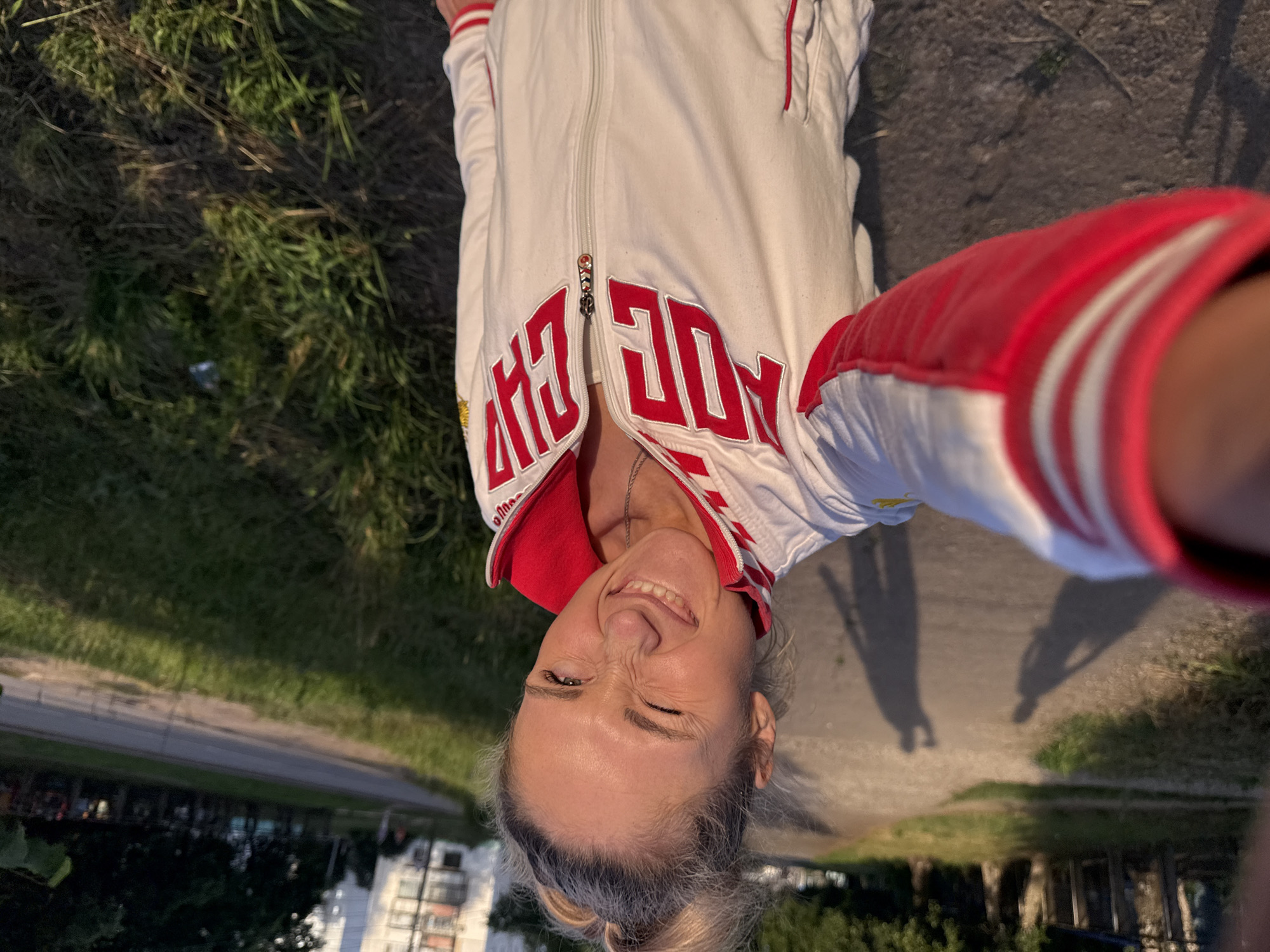 The media file features a self-portrait of a woman outdoors. She is wearing a white jacket with red accents and lettering on the front. The jacket has a zipper and a red collar. The woman has blonde hair, pulled back in a bun, and she is smiling with her eyes squinted, suggesting a playful or happy emotion. She is wearing a silver necklace.
The setting appears to be a sidewalk or paved path next to a grassy area. In the background, there is a road and some buildings, including apartment blocks. The overall impression is of a casual, daytime scene. Shadows are visible on the ground, indicating the presence of sunlight and suggesting it is either morning or late afternoon. The lighting suggests it is daytime. The image is taken at an unusual angle, likely by the subject holding the camera at arm's length.
Visible text on the jacket reads "РОССИЯ", which translates to "RUSSIA". This text is in large, bold, red lettering. There is also a small emblem on the zipper pull. The location context provided is Chaikovsky, Russia, which aligns with the visible text.