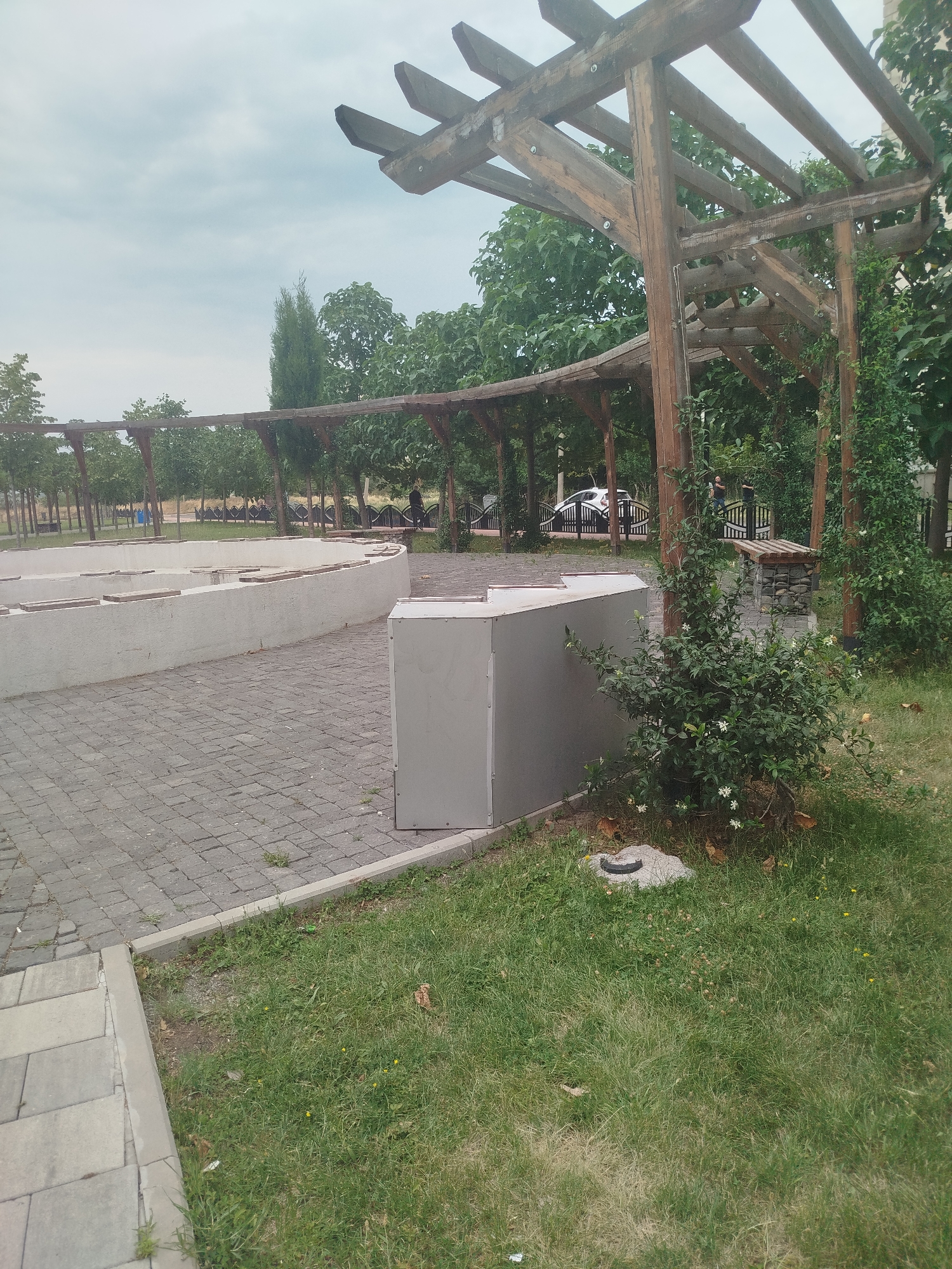 The image depicts an outdoor seating area with a wooden pergola structure in a park-like setting. The pergola, constructed from rough-hewn wooden beams, features a curved roof and supports. In the foreground, a paved area made of grey rectangular stones transitions to a grassy area. A low, curved concrete structure, possibly a planter or seating area, is visible with wooden slats placed on top. A grey rectangular metal bin stands to the right of this structure. Behind the seating area, a line of trees forms a backdrop, and a white car is parked near a black metal fence. The sky is overcast. This scene is located in Rustavi, Georgia.