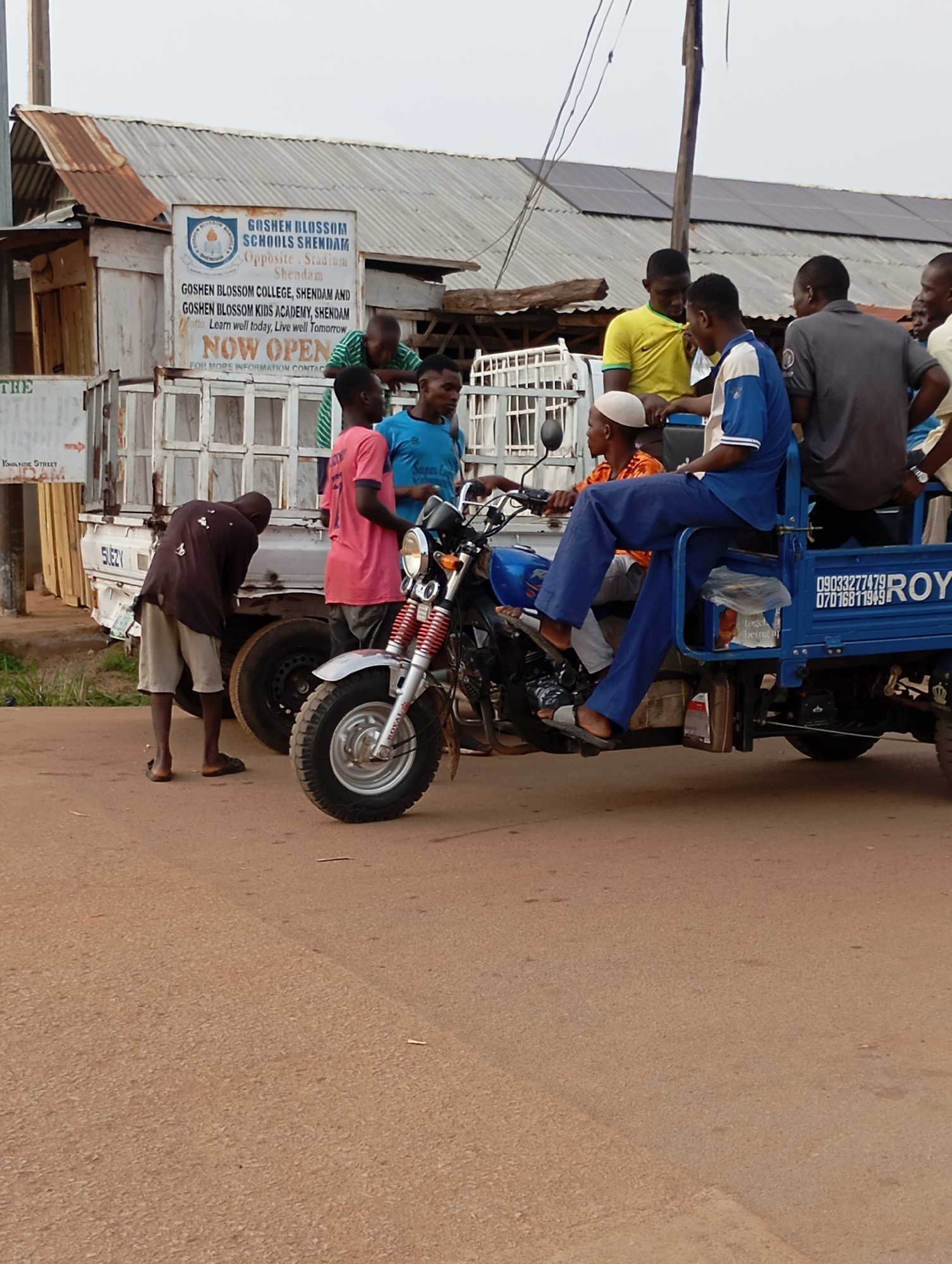In the city of Shendam, Nigeria, a group of individuals are gathered around a three-wheeled motorized vehicle. The vehicle, painted blue, is loaded with goods and has the word "ROYAL" prominently displayed on its side, along with phone numbers: 09033277479 and 07016811949. A man in a dark shirt and light shorts is bending over near the vehicle, possibly making adjustments. On the vehicle's bench seat, a man in a blue shirt and pants is seated, and another man in a pink shirt is standing next to him. A young boy in a green and white striped shirt leans against the vehicle's canopy.
In the background, a building with a corrugated metal roof, partially covered with solar panels, stands adjacent to a smaller wooden structure. A sign on the building reads "GOSHEN BLOSSOM SCHOOLS SHENDAM," with additional text detailing "GOSHEN BLOSSOM COLLEGE, SHENDAM AND GOSHEN BLOSSOM KIDS ACADEMY, SHENDAM." Below this, the words "NOW OPEN" are visible. A white truck is parked behind the three-wheeled vehicle. The ground is paved with asphalt.