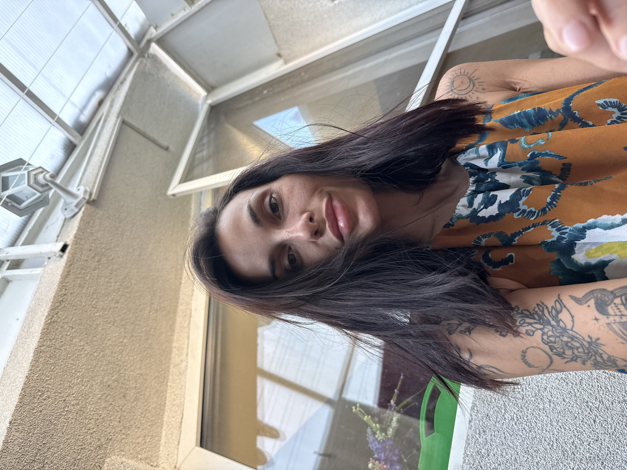 A woman with dark hair is seen in a close-up, partially obscured by her hair. She has a fair complexion and light-colored lips. She is wearing a patterned top with yellow, blue, and white colors. Part of her arm is visible, adorned with tattoos. The background suggests an outdoor or semi-outdoor setting, with an architectural structure, likely a window or enclosure, constructed of white material. There are signs of greenery, such as a plant with flowers, partially visible in the lower part of the image. The image location could potentially be Pleven, Bulgaria, but there are no distinct landmarks to confirm this.