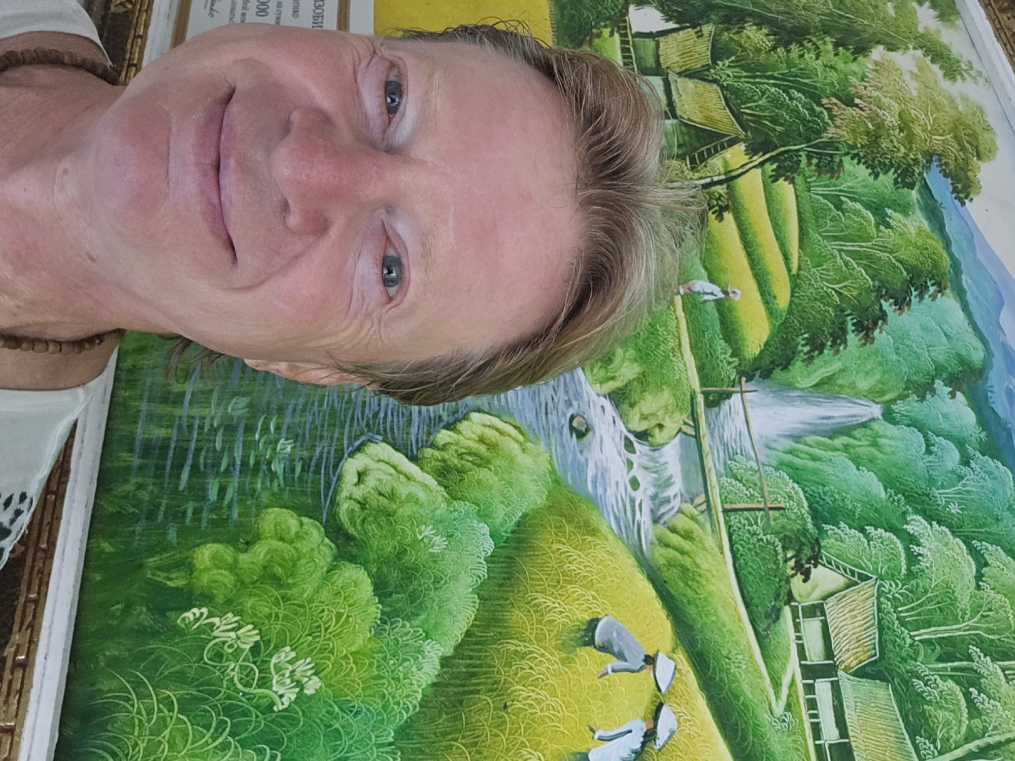 A man with light skin and blondish-brown hair smiles at the camera, positioned in the foreground of a vibrant painting. The painting depicts a lush, green landscape with rice paddies, a waterfall cascading down a cliff, a winding river, traditional houses with thatched roofs, and figures in conical hats working in the fields. The man wears a white shirt and a beaded necklace. The painting appears to be a mural or a large artwork displayed in a public space, potentially an art gallery or a tourist attraction in Denpasar, Indonesia.