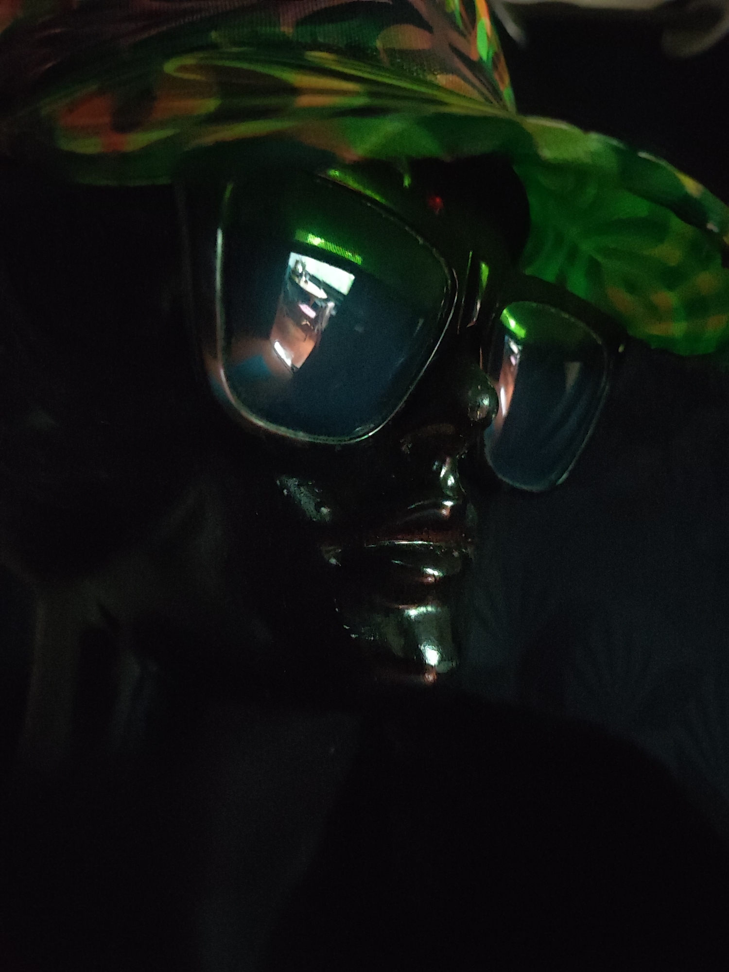 The image shows a close-up of a dark, shiny mannequin head adorned with a colorful hat and dark sunglasses. The hat has a camouflage-like pattern with shades of green, brown, and orange. The sunglasses are large and wrap-around, reflecting a bright, possibly indoor scene with tables and chairs, suggesting an interior setting such as a cafe or restaurant. The mannequin's face is also reflective, showing highlights from lighting. The background is predominantly dark and out of focus, with some faint textures visible. The scene appears to be set indoors, and no specific location in Ko Pha-ngan District, Thailand, can be identified from the visual information alone.
