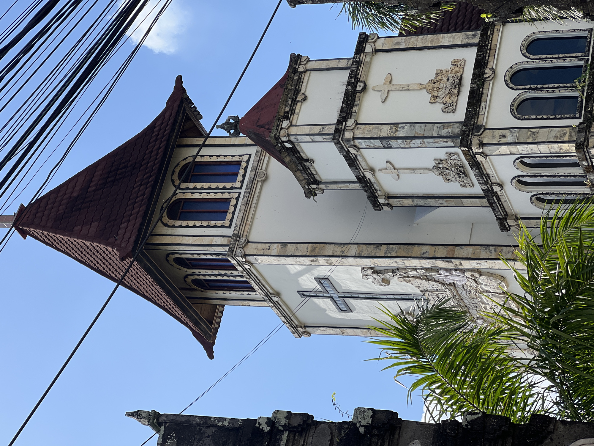 A white church tower with a red tiled roof stands against a clear blue sky, partially obscured by green palm fronds in the foreground and black electrical wires crisscrossing overhead. The tower features multiple levels, each adorned with decorative architectural elements and religious symbols, including crosses. The main tower has arched windows with dark panes and ornate carvings around the facade. A smaller, adjacent tower section mirrors some of the decorative styles. The overall impression is that of a tropical religious building.