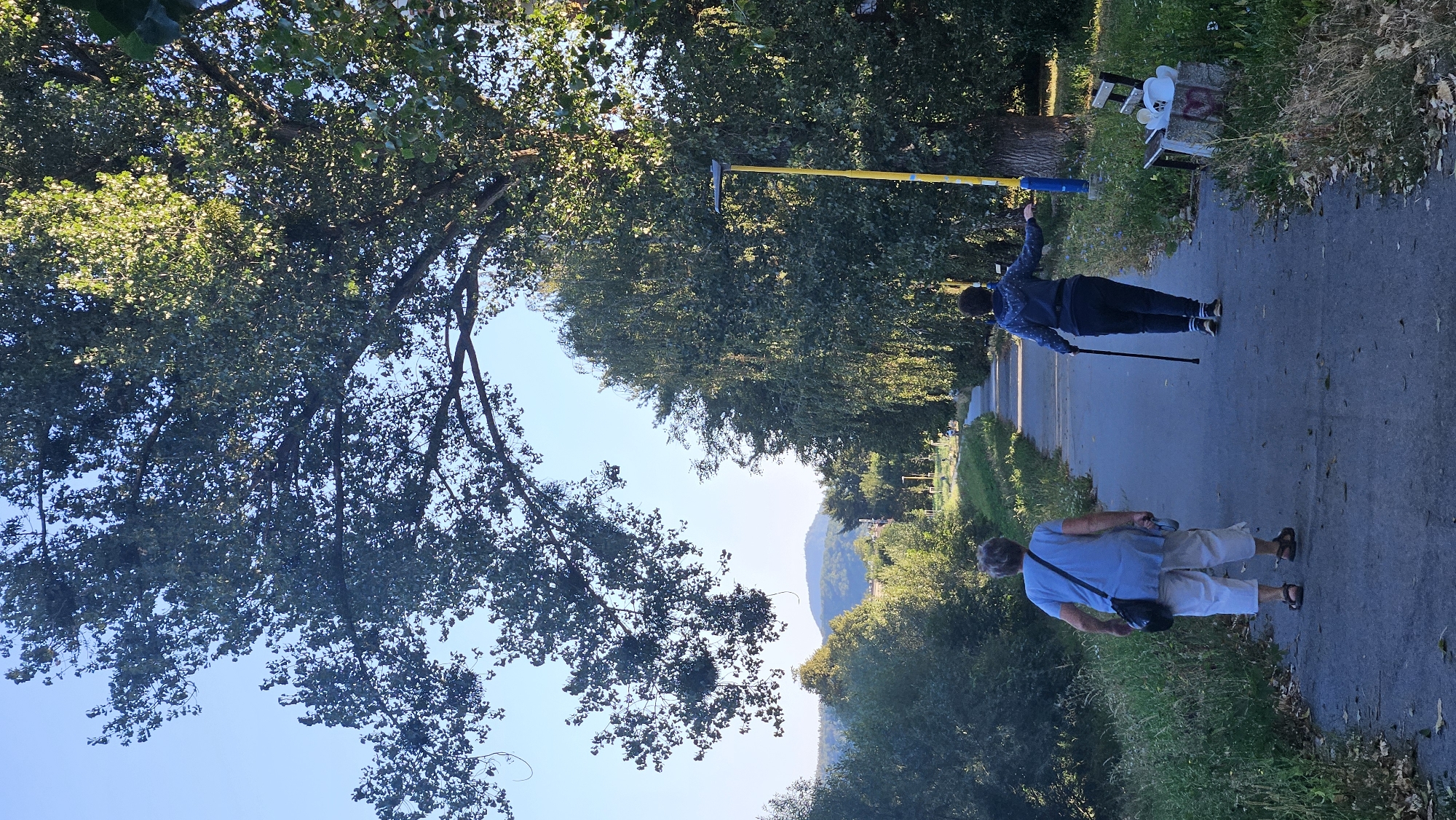 The image shows a tree-lined path in the city of Prešov, Slovakia. Two individuals are walking away from the camera on the path. The person in the foreground on the left is wearing a light blue short-sleeved shirt and white capri pants, carrying a dark bag over their left shoulder and a leash in their right hand. The second person, further ahead on the right, is wearing a dark blue hooded jacket and dark pants, using a walking stick in their right hand and extending their left arm. A lamppost with a yellow pole and blue base stands to the right of the path, near a bench with some objects on it. The path is bordered by green grass and vegetation, with a line of trees forming a canopy overhead and along the right side of the path. In the distance, rolling hills are visible under a clear blue sky. The scene appears to be a park or a riverside promenade.