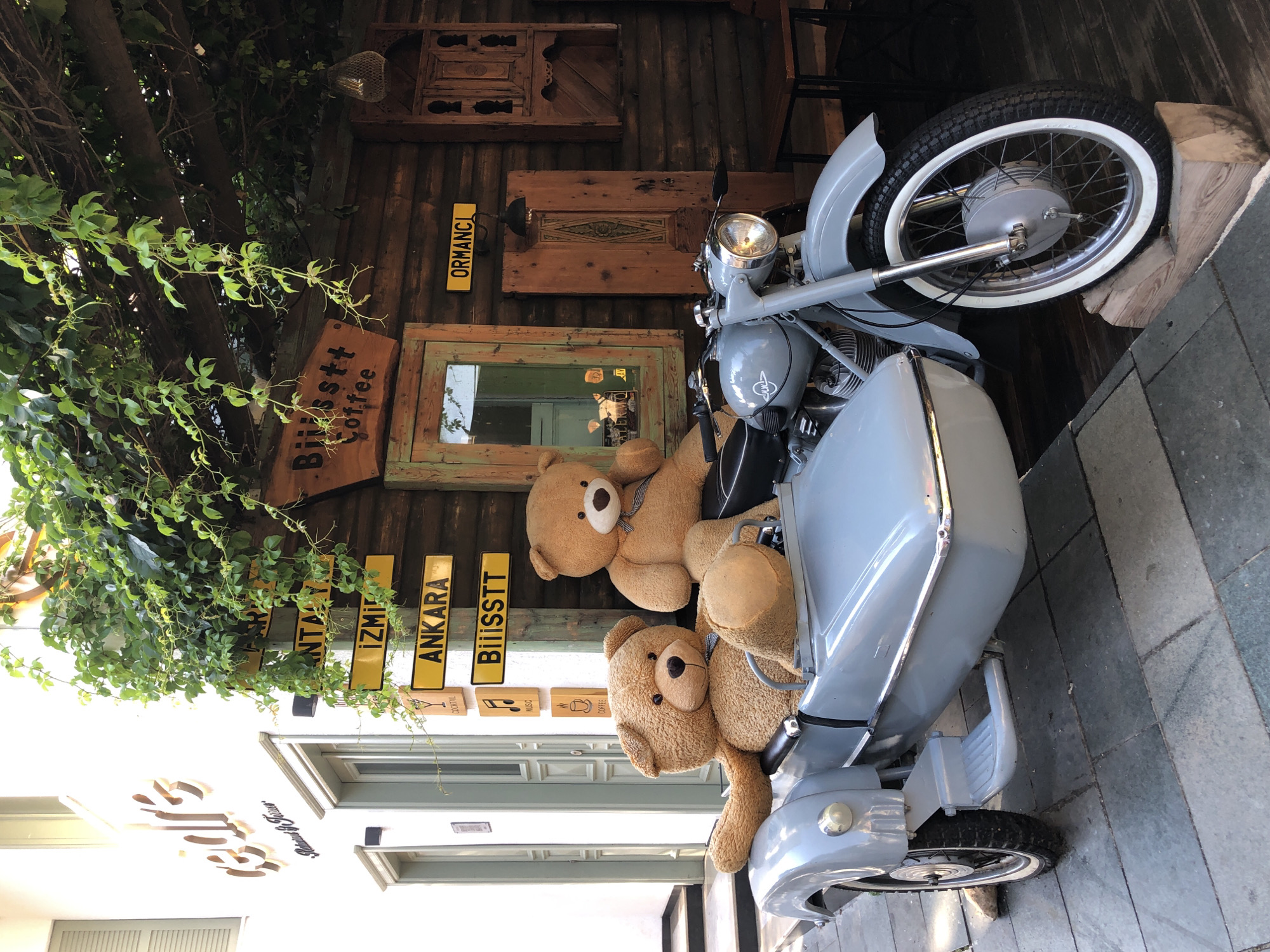 In Kaş, Turkey, a light blue motorcycle with a sidecar is parked on a cobblestone pathway. Two large teddy bears are seated in the sidecar. The motorcycle is positioned in front of a rustic wooden building, which appears to be a cafe or shop, as indicated by signage that reads "Biiisss tt Coffee" and directional signs pointing to "ANTALYA," "İZMİR," "ANKARA," and "ORMANCI." Ivy vines hang from an overhead wooden pergola, partially obscuring the building. To the left of the building is a white structure with a sign that says "cedre." The scene is outdoors, under natural light.
