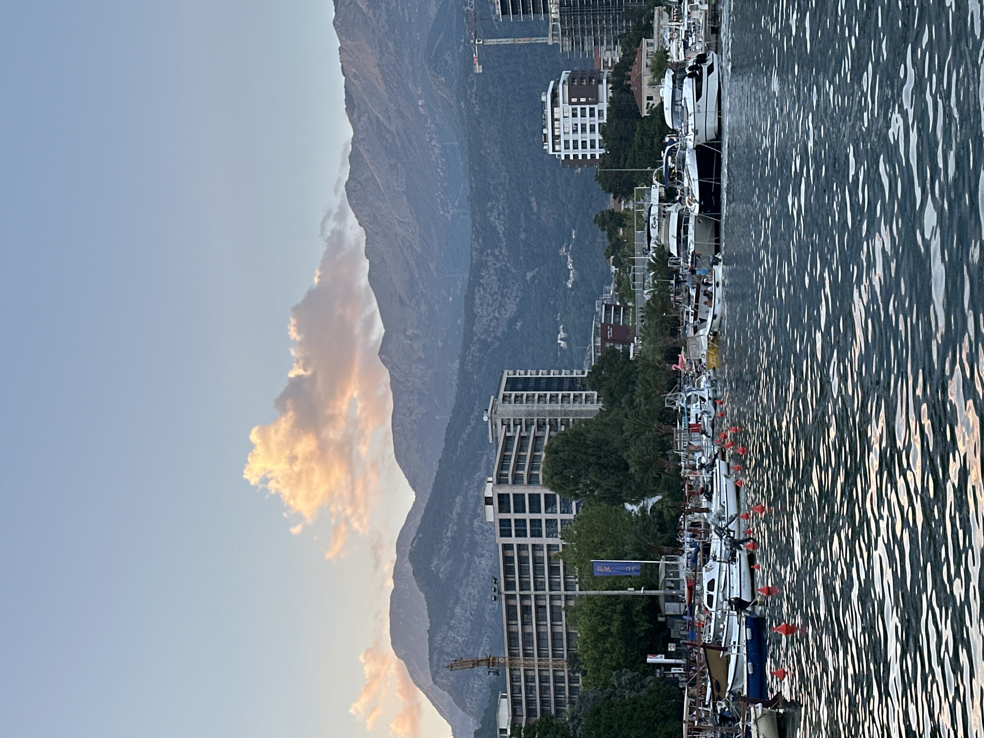 The image depicts a harbor in Budva, Montenegro, at sunset. The foreground is dominated by a body of water with choppy waves, reflecting the sky and surrounding structures. A line of small, orange buoys marks a boundary within the water. Moored in the middle ground are numerous boats, including several larger yachts with white hulls and dark accents, as well as smaller fishing boats. Some boats have people on board.
Behind the harbor, a cityscape unfolds with several modern buildings. One prominent building is a long, multi-story structure with a beige facade and numerous windows, alongside which a construction crane stands. Another building, appearing more residential, has a white facade with dark window frames. Further buildings, some under construction, are visible.
In the background, a range of rugged mountains forms the horizon. The sky is a soft gradient of blue and pink, with a single, large, fluffy cloud illuminated by the setting sun. Trees, including palm trees, are interspersed with the buildings, adding a touch of greenery to the urban landscape. The overall atmosphere is serene, indicative of early evening.