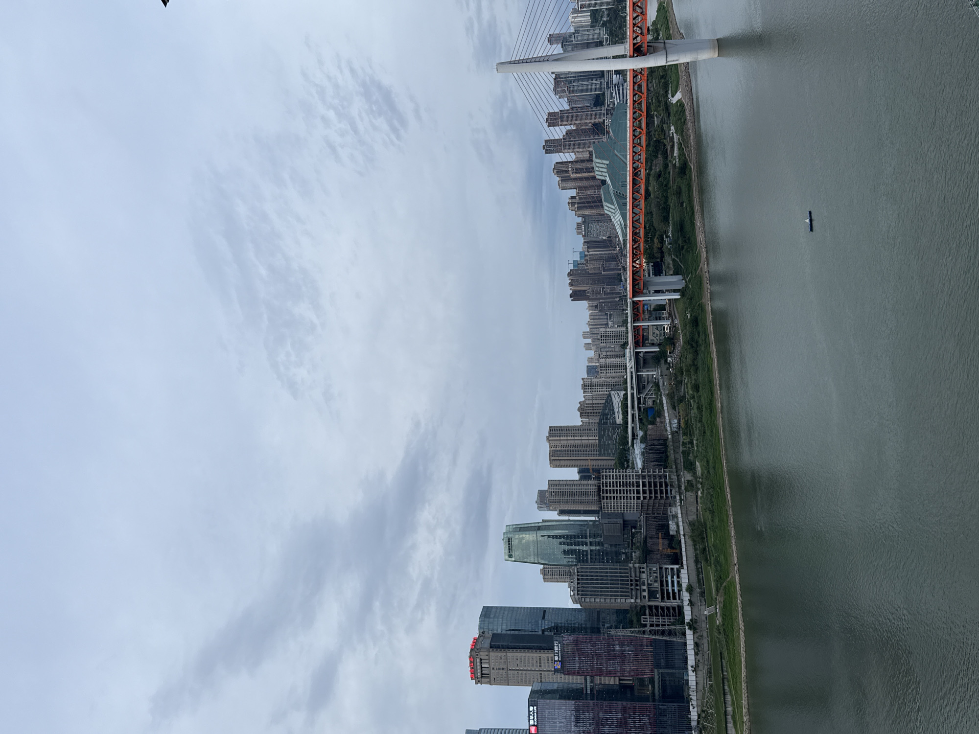 The image depicts an aerial view of the city of Yuzhong District, China, showcasing a skyline dominated by high-rise buildings along the riverfront. The river flows vertically on the right side of the frame. A suspension bridge, painted with orange accents, spans the river with cables connecting to a central white tower. A single vessel is present in the river. The sky is overcast with grey clouds.