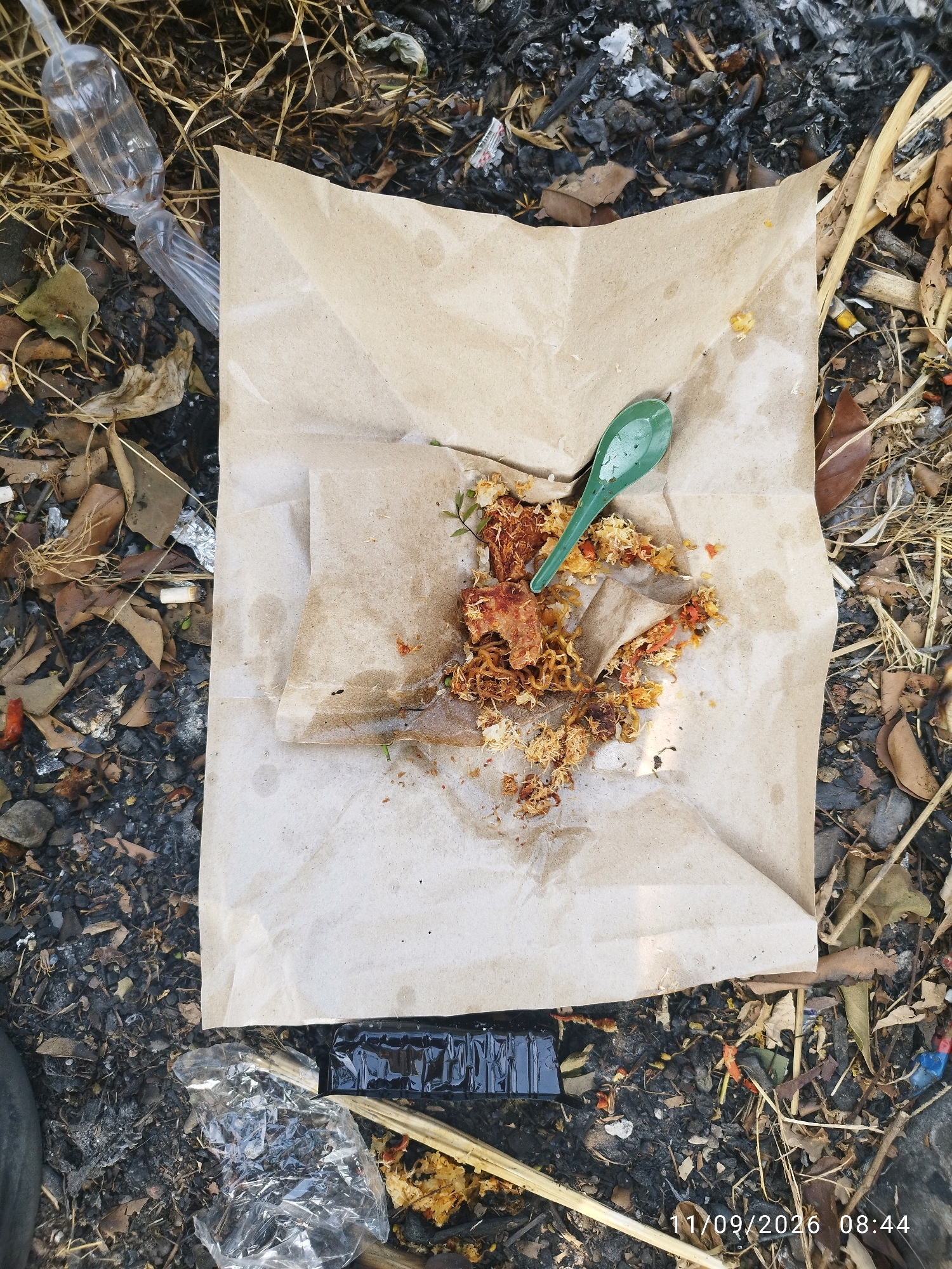 The image shows a top-down view of a paper-wrapped food parcel and a green plastic spoon lying on a surface covered with dry leaves, twigs, and debris. Some of the food appears to have spilled onto the paper wrapper and the surrounding ground. To the left, there is a clear plastic bottle. In the lower section of the image, there are pieces of clear plastic and a black rectangular object. The background appears to be a mix of dry organic matter and what might be ashes or burnt material. A timestamp "11/09/2026 08:44" is visible in the bottom right corner. The scene suggests a discarded food parcel in an outdoor, possibly unkempt, environment. The location in the city of Mojokerto, Indonesia, implies this scene is in an urban or peri-urban setting. There are no people visible in the image.