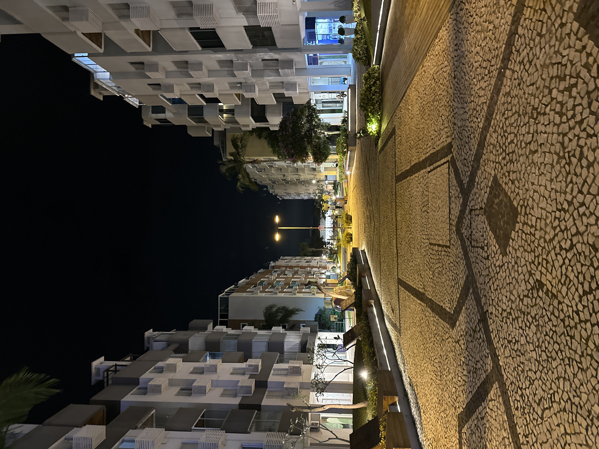 The image depicts a wide pedestrian walkway paved with irregular light-colored stones, featuring a dark geometric inlay pattern. The walkway is bordered by a modern landscaped area with seating and illuminated by linear lights. On either side of the walkway are multi-story apartment buildings with balconies. The buildings are contemporary in design, featuring white facades with grey accents and horizontal louvers on the balconies. The scene is illuminated by streetlights and architectural lighting, indicating it is nighttime. In the distance, several people are walking along the path and some vehicles are visible. The environment suggests an urban or residential complex setting. Specifically, this location is the Passeio Pedra Branca in Palhoça, a municipality within the metropolitan region of Florianópolis, Brazil.