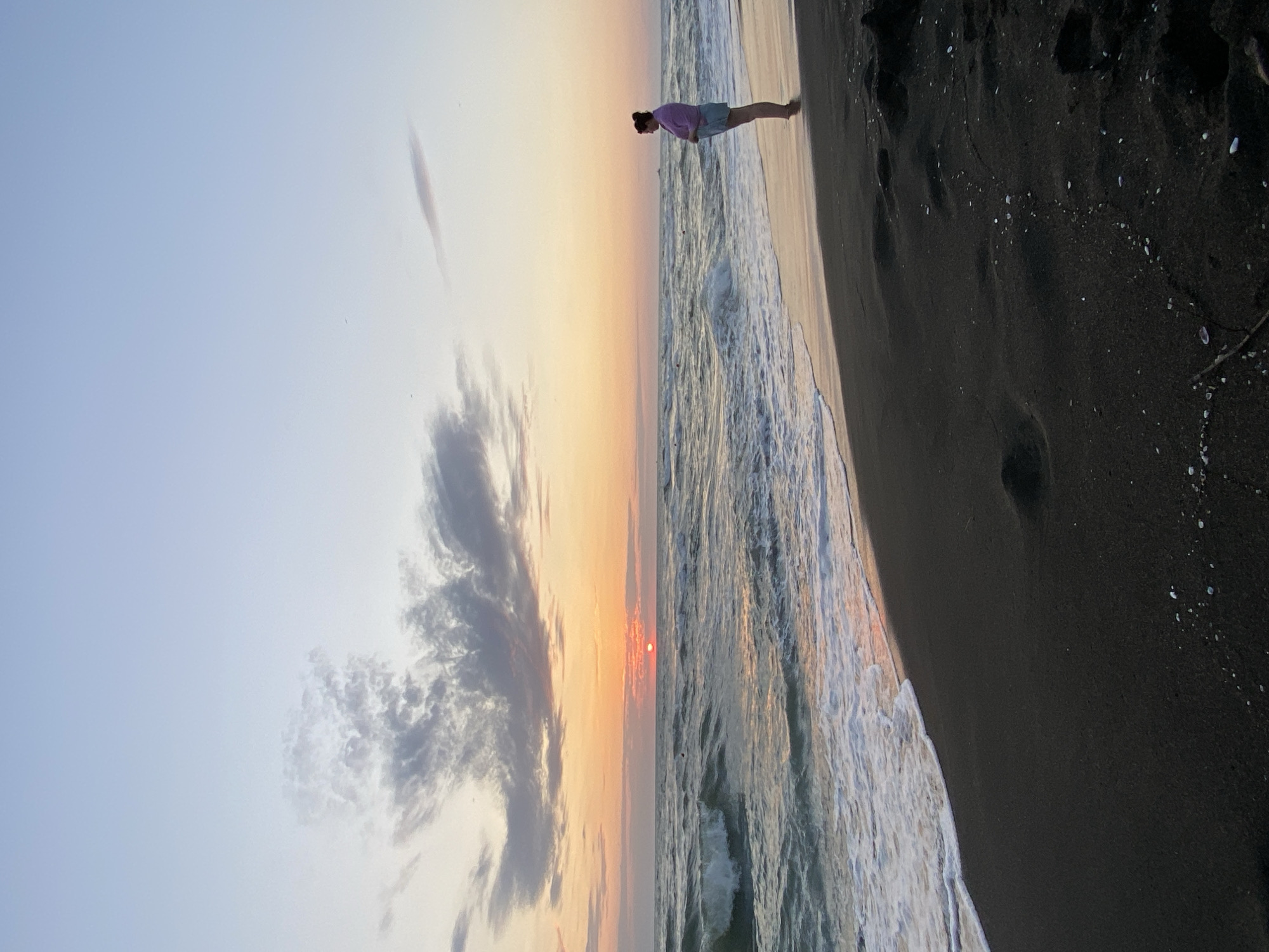 An individual stands on a black sand beach in Ureki, Georgia, facing the sea at sunset. The sun, a small, bright orb, is low on the horizon, casting an orange glow across the sky and reflecting on the water. Waves gently lap the shore, creating white foam. The sky is a gradient of pale blue to soft orange, with scattered clouds. The person is dressed in a light purple top and denim shorts, with their hair pulled back. They appear to be looking out at the ocean. The scene is peaceful and serene.