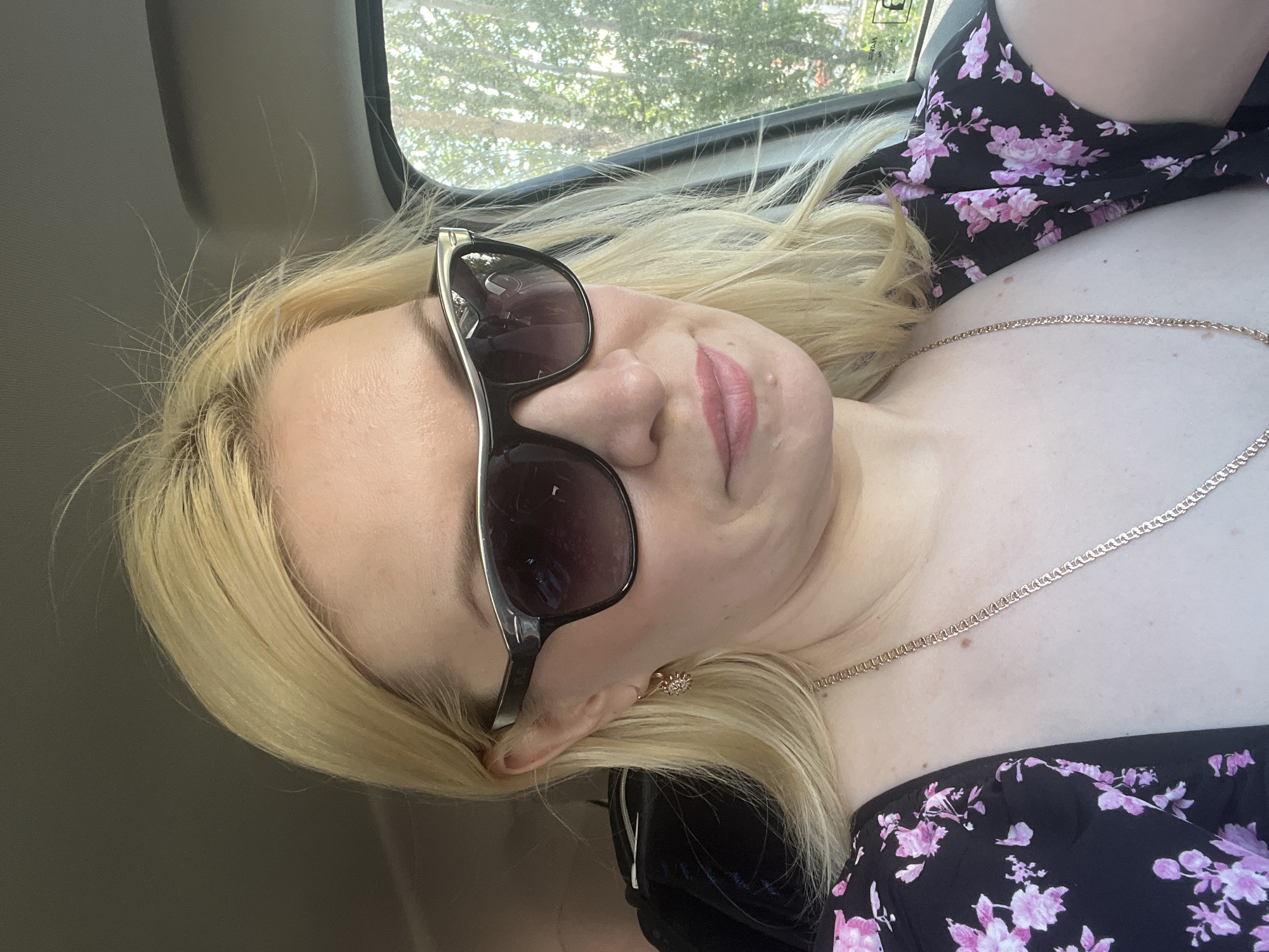 A blonde woman wearing sunglasses and a black floral dress is sitting in a car. She is looking down and to the side with a slight smile. The background shows trees and greenery, indicating an outdoor environment. The scene appears to be a selfie taken inside a vehicle. The woman is wearing a gold chain necklace and a small, intricate earring.