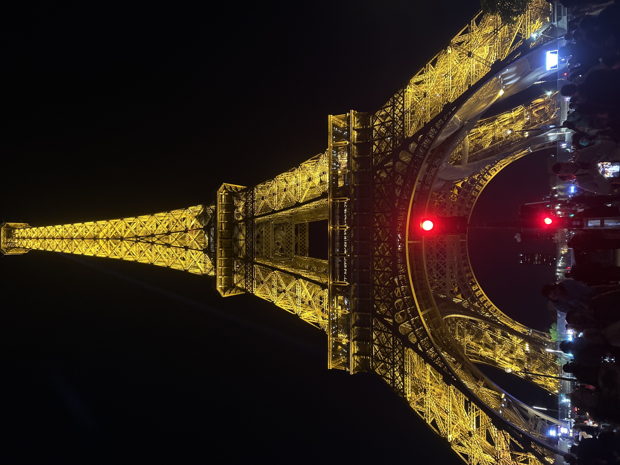 The image shows the Eiffel Tower at night in Paris, France. The tower is lit up with yellow lights and there are two red lights on the lower levels. A crowd of people are observing the tower. The sky is dark and clear. The image is oriented sideways.
