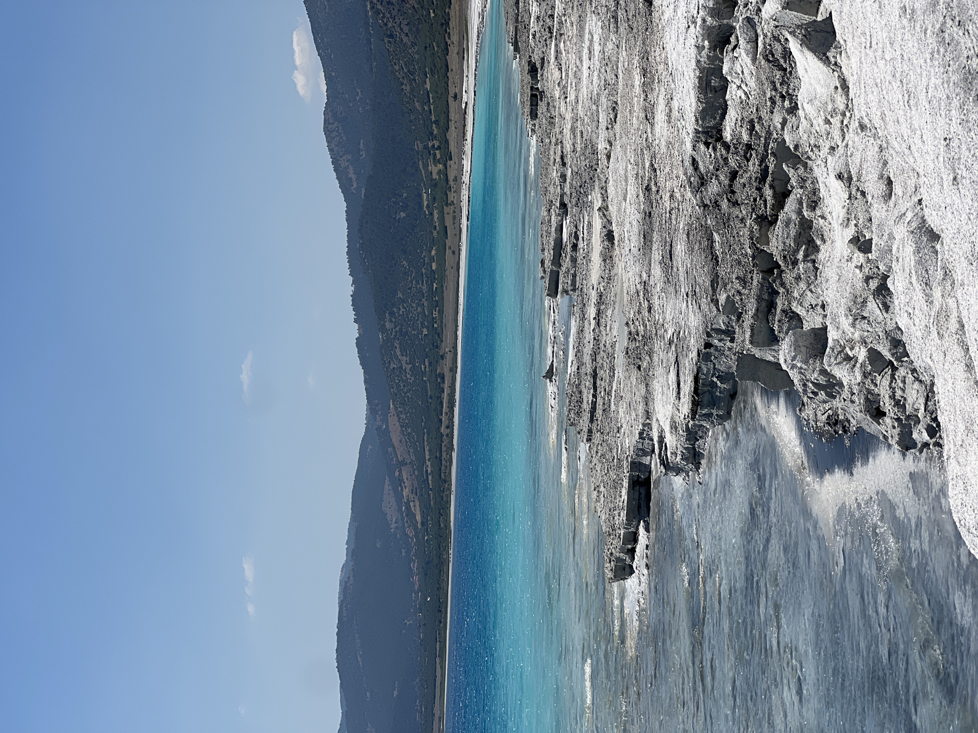 This is a vertical, eye-level, outdoor photograph of Salda Lake in Yeşilova, Turkey. The foreground is dominated by white, crystalline mineral deposits along the shoreline, with some of these deposits appearing as layered rock formations. The lake itself is a vivid turquoise blue, with a slight ripple on the surface reflecting the bright sunlight. The water appears shallow and clear near the shore, gradually deepening to a more intense blue further out. In the mid-ground, the opposite shore of the lake is visible, lined with a thin strip of sandy beach and then a gentle slope covered with sparse vegetation and scattered trees. The background consists of rolling, treeless hills that rise to form a mountainous landscape, with a clear, pale blue sky overhead. There are no people or animals visible in the frame. The overall impression is one of a pristine natural landscape under a bright, sunny day.