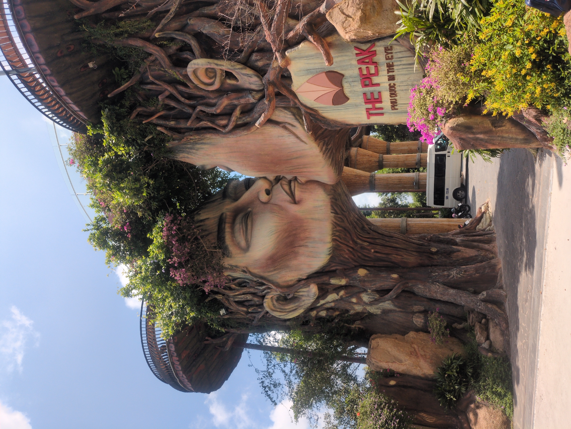 The image depicts an artistic wood sculpture in Phú Quốc, Vietnam, representing two faces in a kissing pose, with hair formed from tree branches and a sign that reads "The Peak Phu Quoc in the eye". The sculpture is decorated with greenery and flowers. A bus is seen parked on the road behind the sculpture. A balcony-like structure is visible in the background on the left side of the image. The sky is visible between the structures and trees.