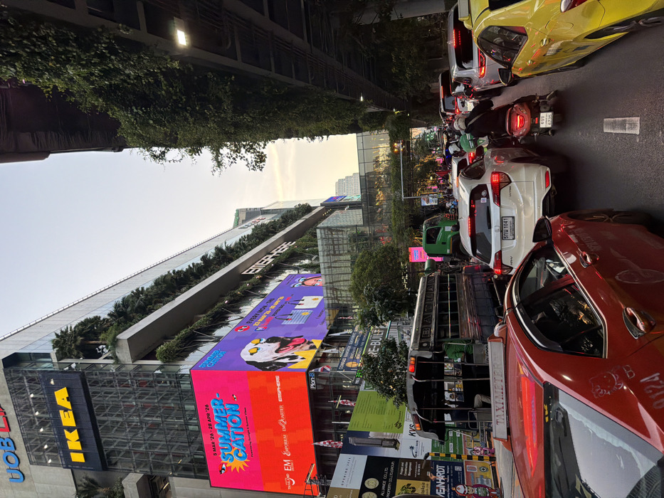 The image depicts a congested urban street scene in Bangkok, Thailand, specifically near the EmSphere shopping complex on Sukhumvit Road. The time of day appears to be late afternoon or early evening, indicated by the fading natural light in the sky.
On the left, a large, modern multi-story building dominates the background. It features prominent "UOB" and "IKEA" signage near its upper sections. Below these, multiple large digital billboards display vibrant advertisements; one features a "SUMMER-CATION" campaign with "EMSPHERE" branding, showing a dog wearing sunglasses. The building's facade incorporates numerous levels of vertical greenery, including palm trees.
To the right, an elevated concrete structure, likely part of an overhead walkway or transport system, is heavily covered in dense green foliage, obscuring its structural details.
The foreground and middle ground are filled with multiple lanes of traffic, appearing stationary or moving very slowly. Visible vehicles include:
* In the immediate foreground, the rear of a red Toyota Corolla Altis taxi, marked "TAXIMETER" on its roof and featuring a "Hello Kitty" sticker on its bumper.
* Behind the red taxi, a dark blue *songthaew* (modified pickup truck with bench seating) is present, with "BATTAN" visible on its side. Several passengers are seated within the vehicle.
* A white Honda HR-V SUV is positioned in the center, displaying a Thai license plate "5 กณ 1041".
* To the right of the white SUV, a person wearing a dark jacket and helmet is riding a red scooter with license plate "699 6735".
* Further into the frame, a yellow taxi is partially visible on the far right, and a green utility truck is visible behind the *songthaew*.
* Multiple vehicles beyond these are visible, with illuminated red brake lights indicating stopped traffic.
The overall scene is characteristic of a bustling urban environment experiencing traffic congestion.