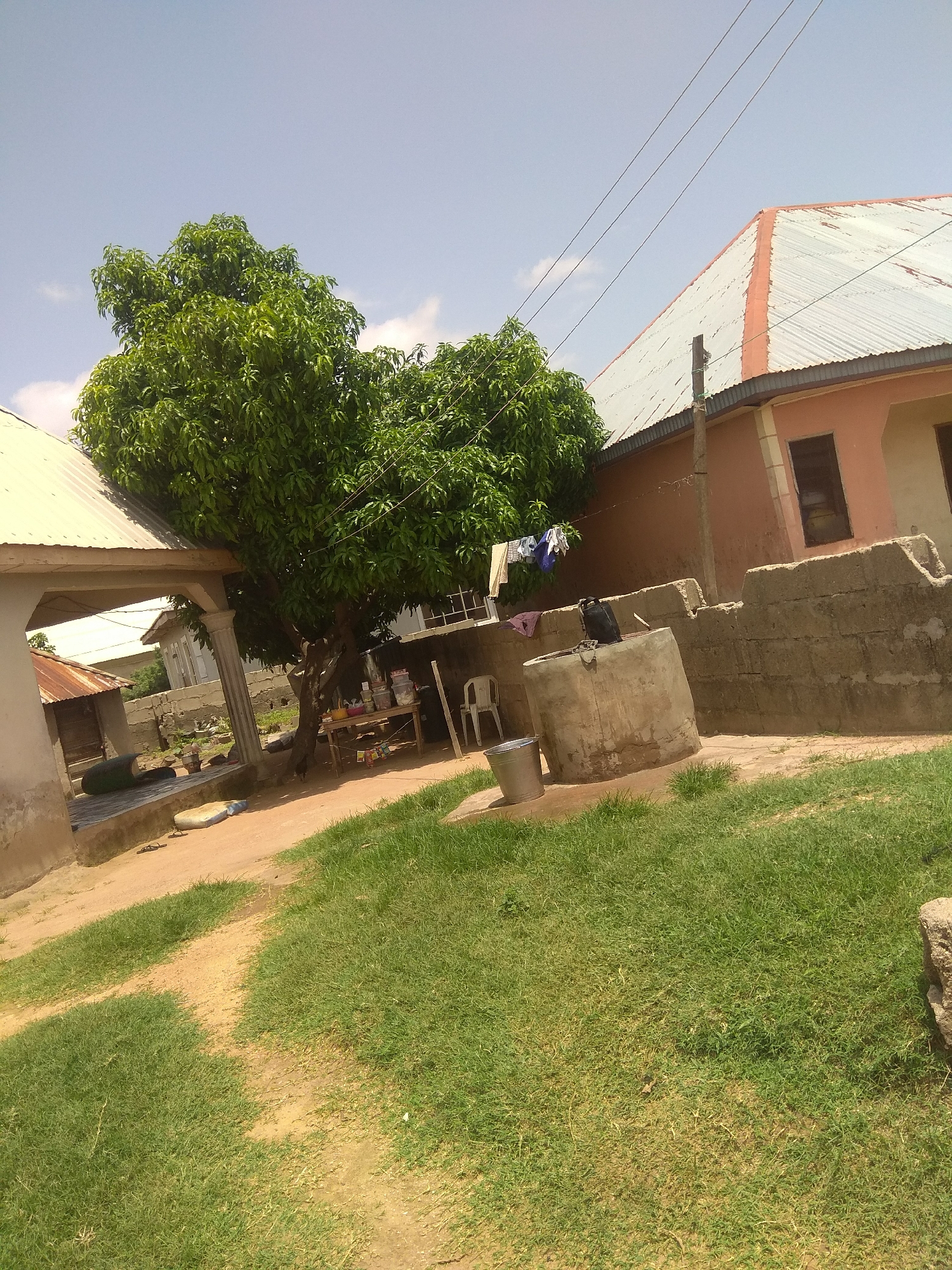 This media file captures an outdoor scene in Jalingo, Nigeria, under a clear, bright sky, suggesting daytime with fair weather. The foreground is dominated by a grassy area with a dirt path winding through it. In the mid-ground, a large, leafy tree stands prominently, its branches offering shade. Beneath the tree, a makeshift table is laden with various items, possibly for sale or personal use. Nearby, a concrete well with a black bag on top is situated, and a metal bucket sits on the ground next to it. A white plastic chair is also visible. Clothes are hanging on a line strung between poles.
Several structures are present, including a building with a corrugated metal roof painted orange and white, appearing somewhat weathered. Another structure with a similar roof is partially visible to the left. A low, rough concrete wall encloses part of the area, with exposed rebar at the top. A tall, thin concrete pillar supports an overhang of one of the buildings. The overall impression is of a residential or commercial area, possibly a market stall or a home with open space. There are no people visible in the image. The scene appears tranquil and everyday.