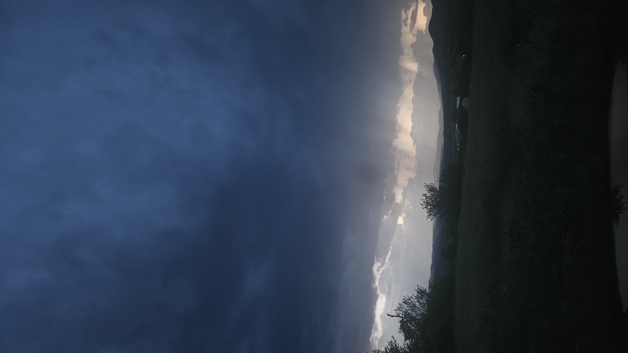 The image depicts a dark, stormy sky with heavy, blue-gray clouds covering most of the frame. Rays of light pierce through the clouds in the distance, suggesting a rain shower occurring over a landscape of rolling hills. The foreground is a dark, silhouetted field of grass, with the tops of some trees visible on the left. The overall impression is one of an approaching or receding storm.